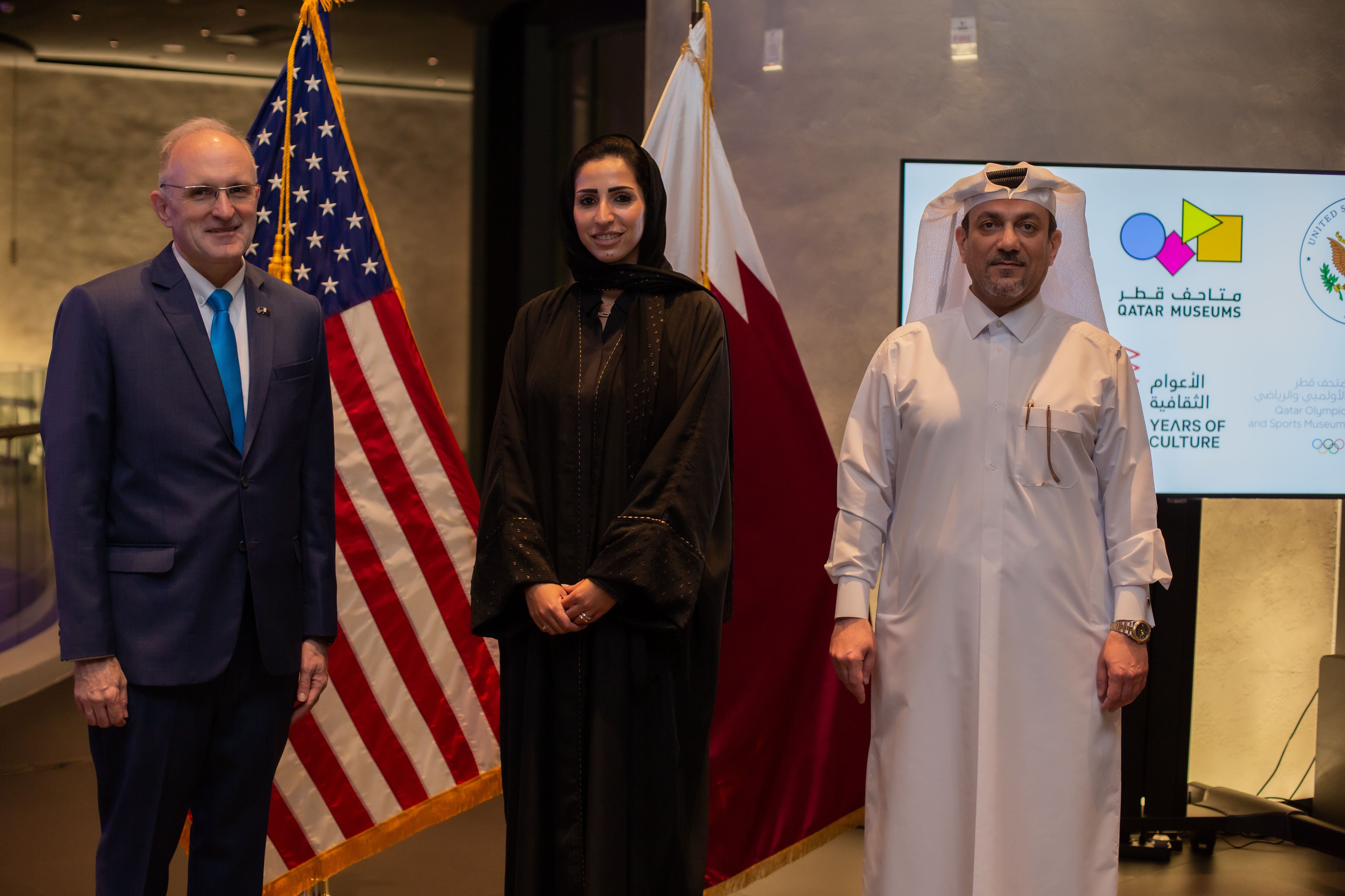 A man in a blue suit beside a man and woman in Qatari clothing, standing and smiling in front of the US and Qatar flags.