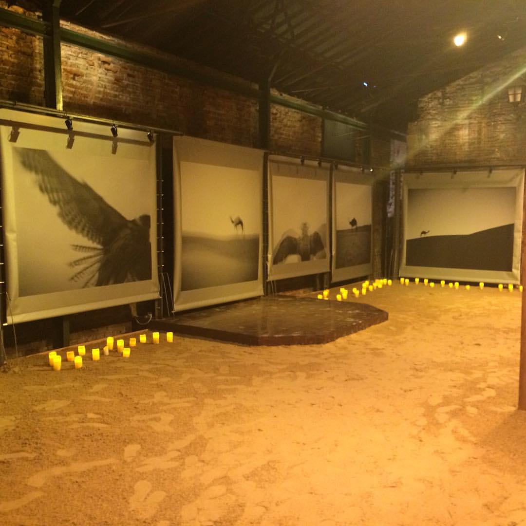 A gallery space with large black and white photography prints on the walls, lit candles candles and sand on the ground.