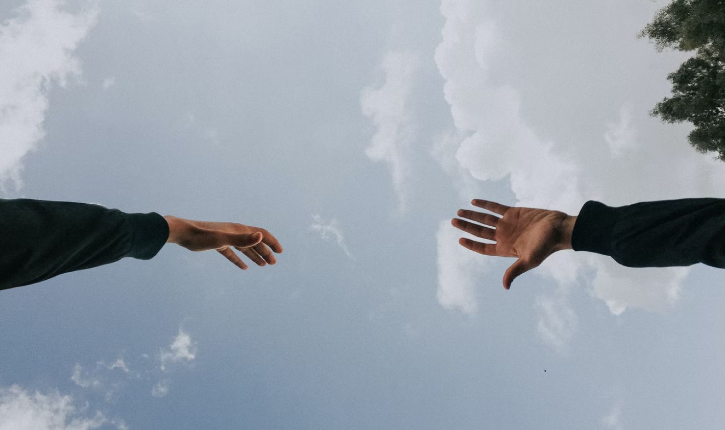 Two people's hands reaching out towards each other in friendship from opposite sides, seen from below against a blue sky.Two people's hands reaching out towards each other in friendship from opposite sides, seen from below against a blue sky.
