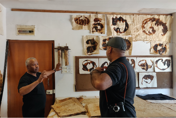 2 men in artist Faraj Daham's studio surrounded by painted artworks, during Qatar-India 2019 Year of Culture.