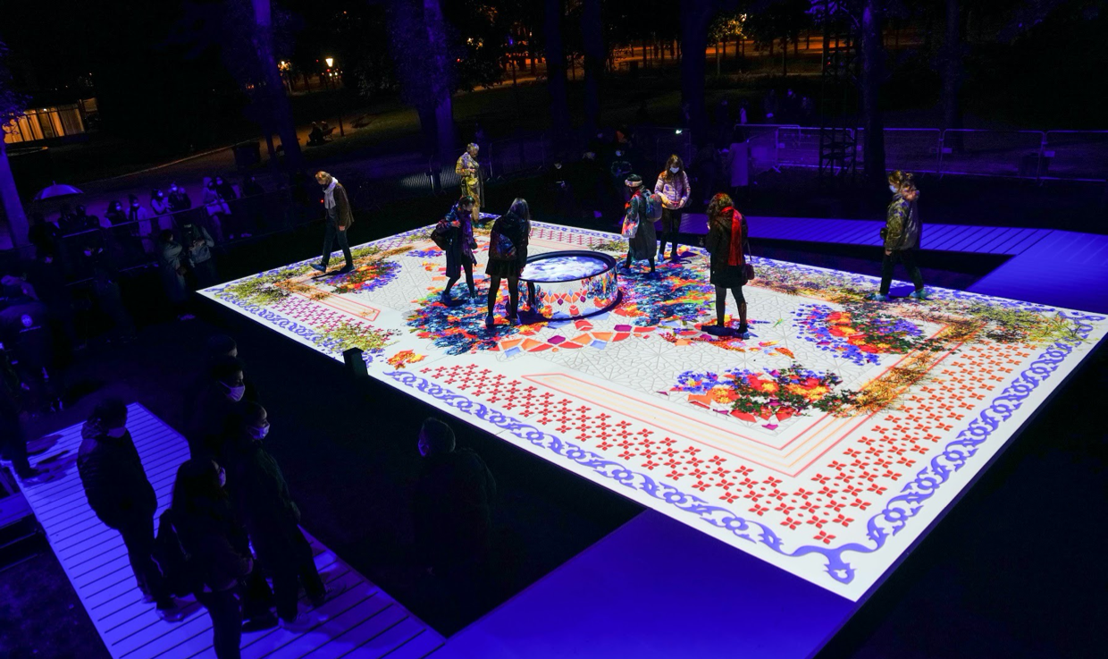 People walk across an interactive digital artwork designed to resemble a Persian rug at a contemporary arts event in Paris.