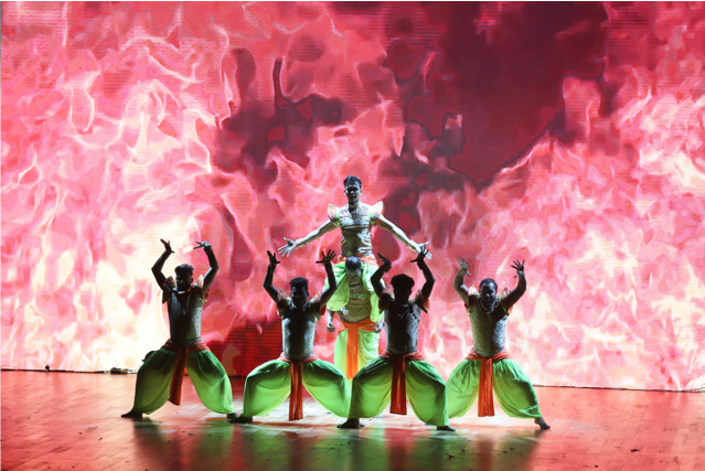 Four male Indian dancers perform Ticket to Bollywood on stage, in front of a red flame backdrop.