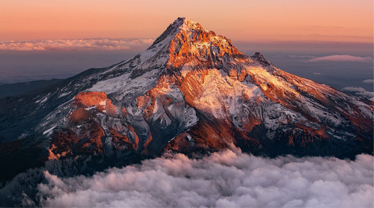 A snowcapped mountain in the Pacific Northwest of the USA, with the orange glow of sunset against an orange sky. 