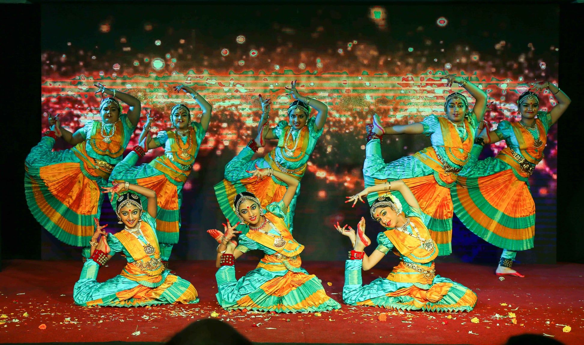 Eight Indian dancers pose on stage wearing orange and turquoise dresses, during the Passage to India event.