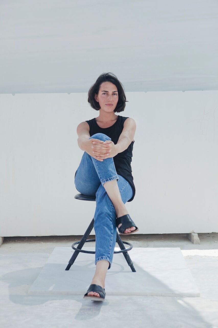 Woman with short dark hair wearing a black tank top and denim jeans sits cross legged on a stool against a white background.