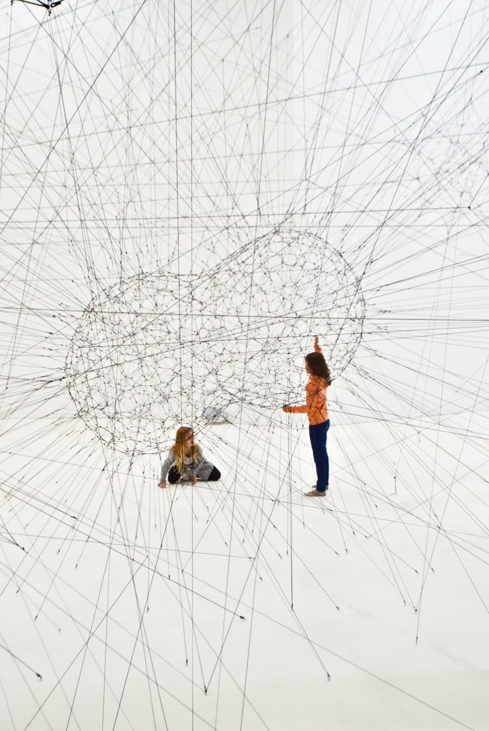 Two children from different cultures play together inside an art installation made of criss-crossing threads.