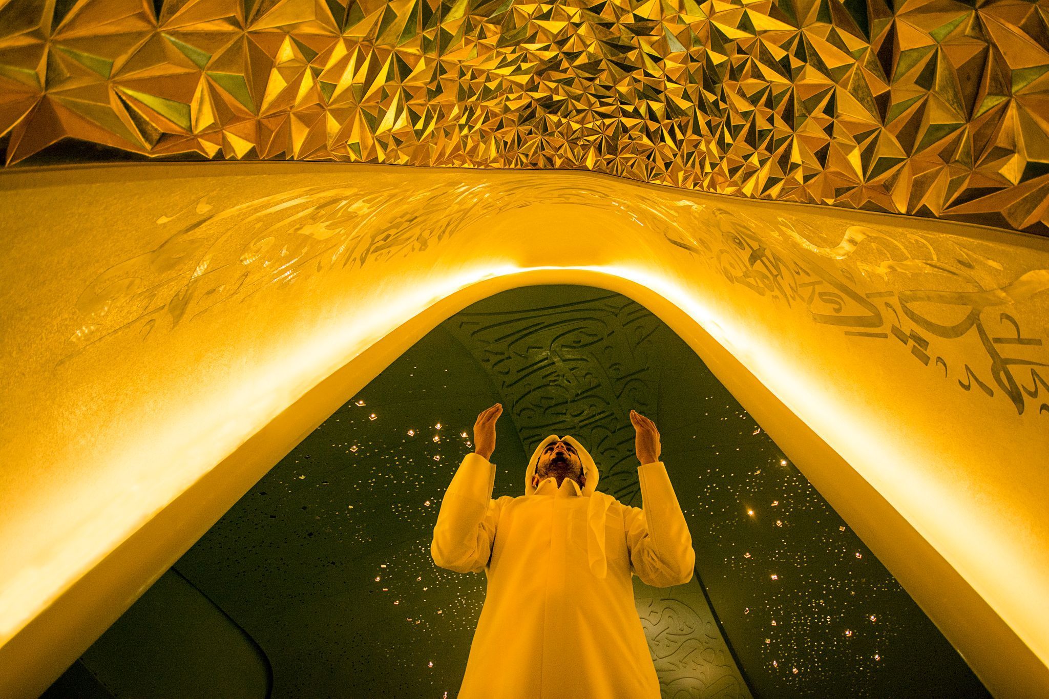 A man wearing all white stands under an illuminated golden archway, with his palms facing upwards during prayer. 