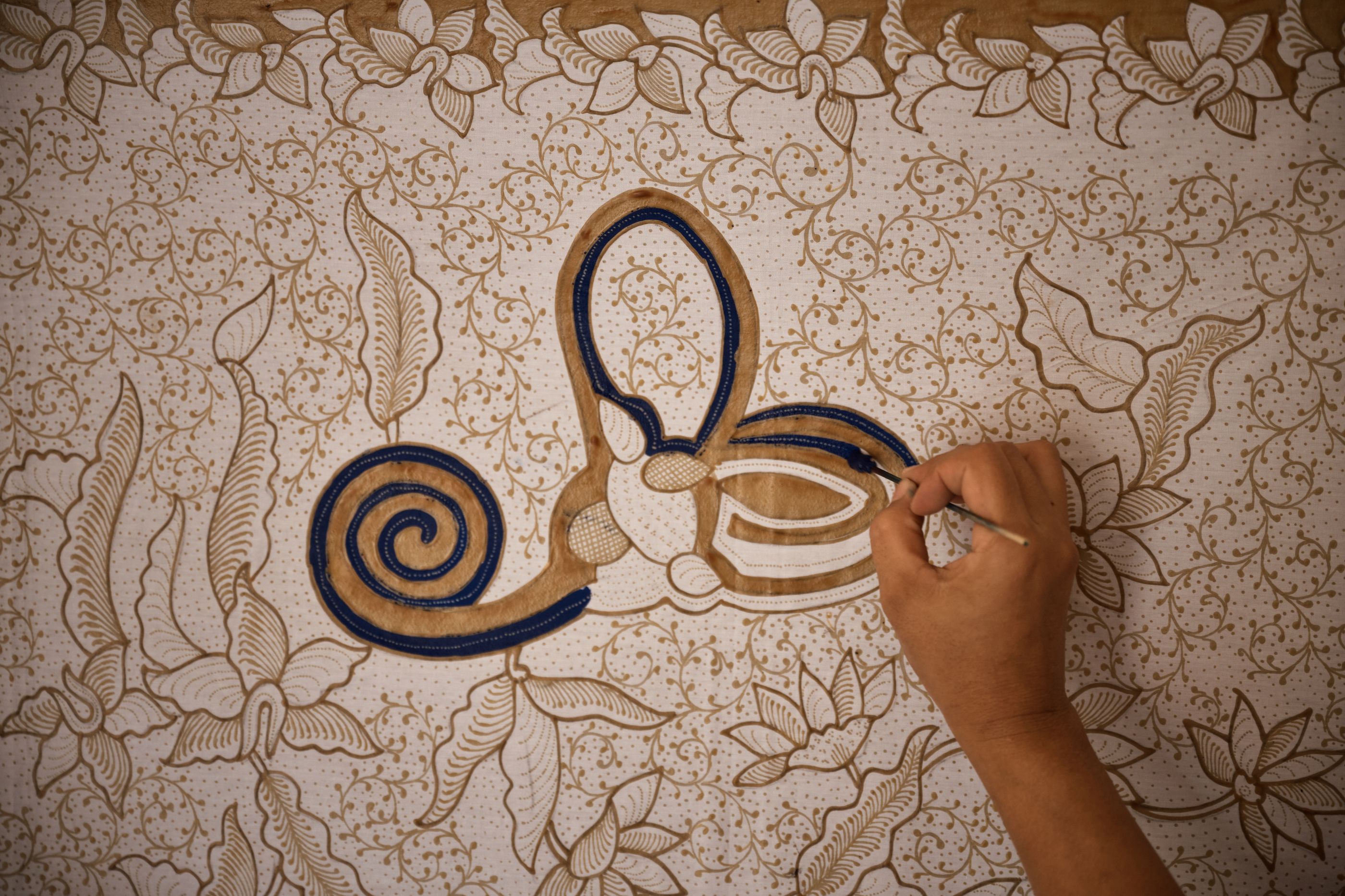 A hand seen from above, painting a traditional Indonesian Batik design onto fabric using brown and black inks.