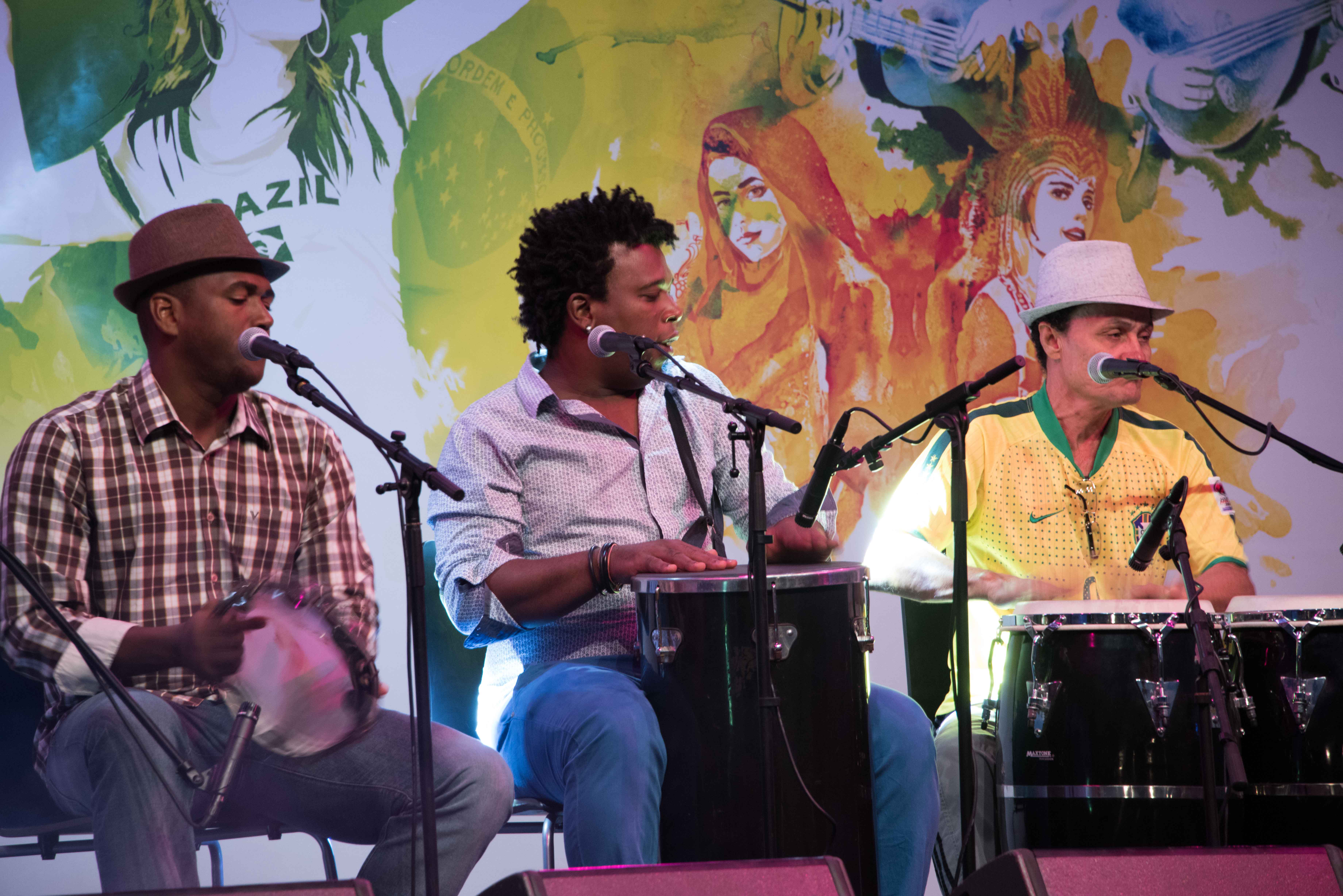 Three men seated on stage playing drums, with a colourful background during the Brazil festival in Qatar.