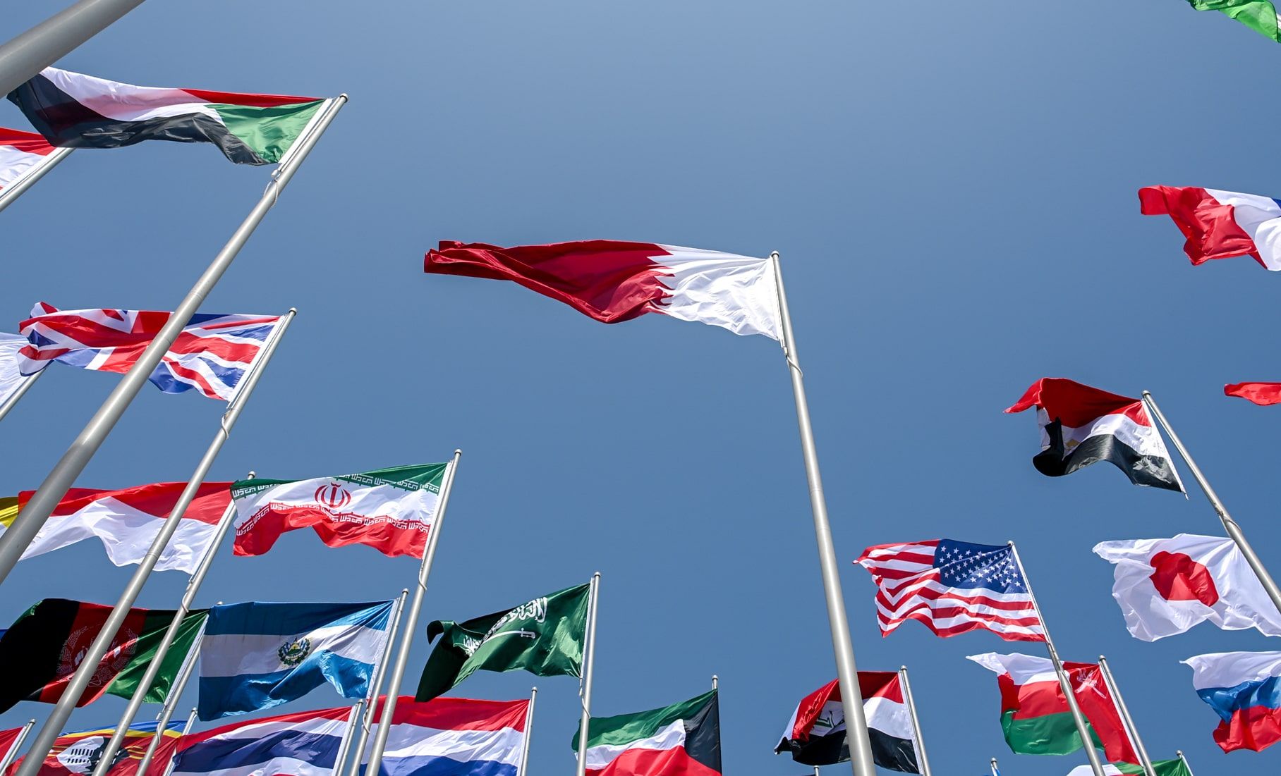 Over twenty international flags flying on flagpoles, seen from below against a blue sky with the Qatari flag at the centre. 