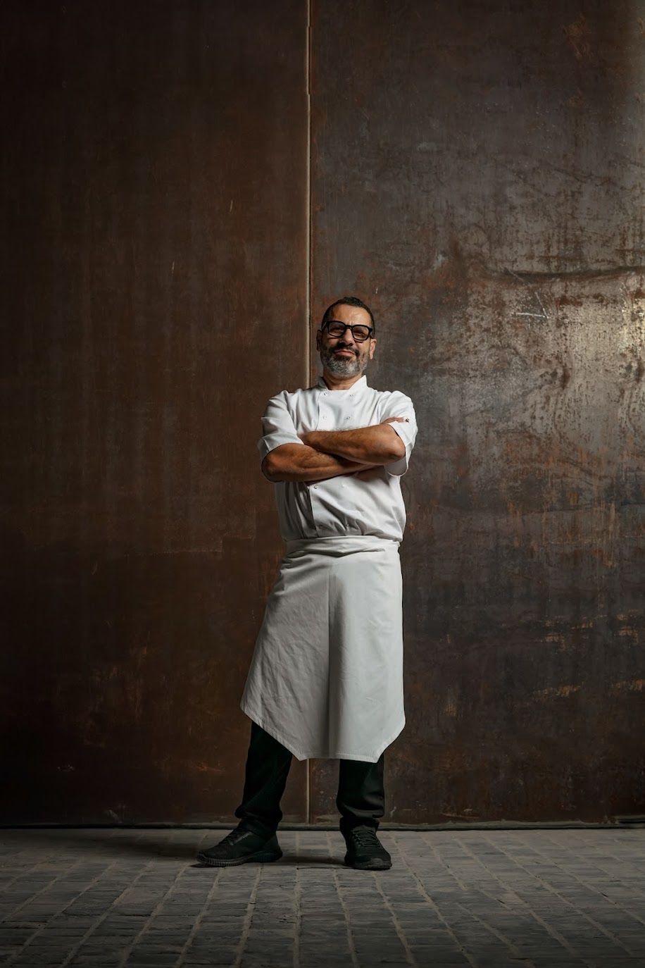 Man wearing glasses, chef whites and apron stands with arms folded and a relaxed neutral expression against a metal wall.