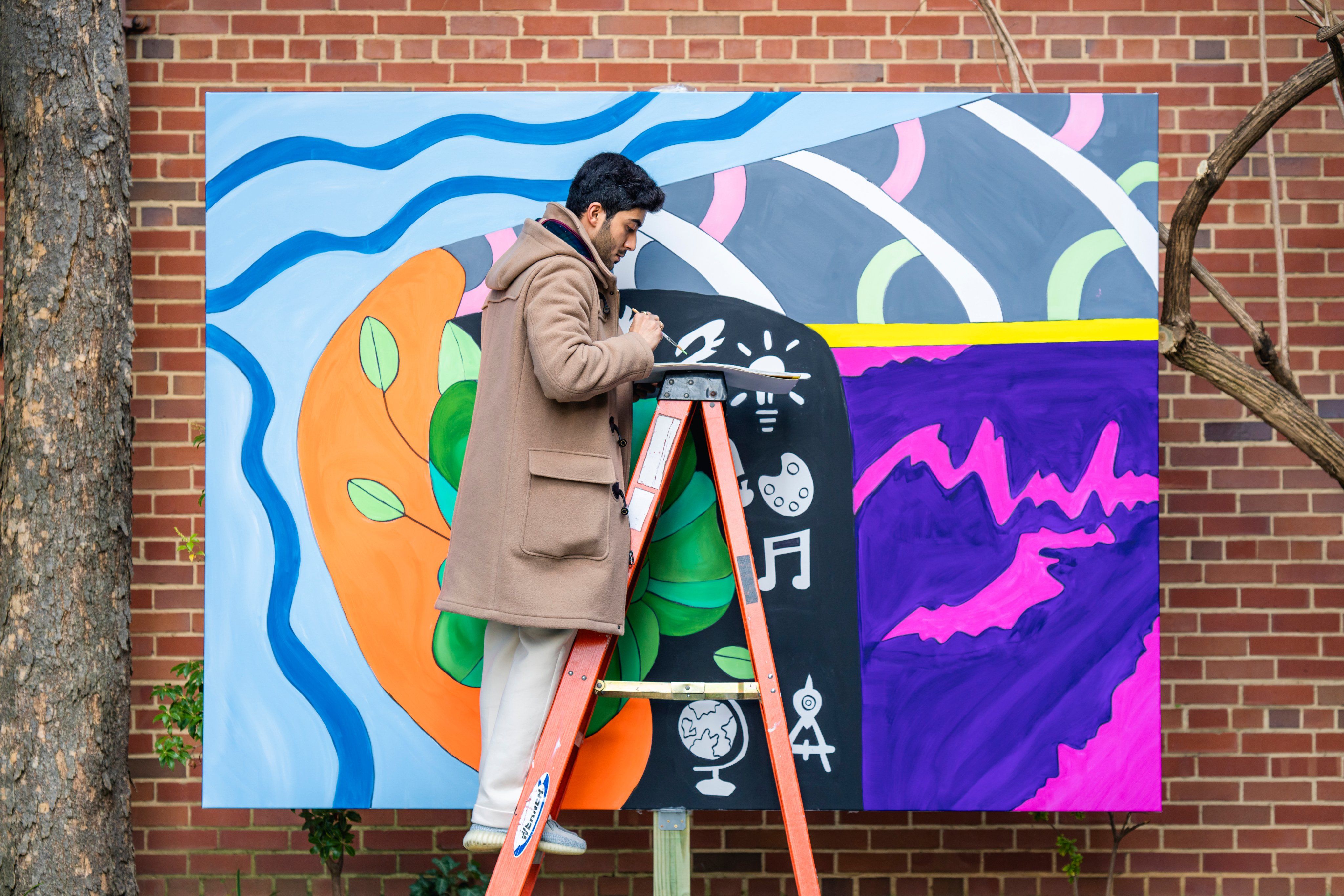 Qatari artist Ahmed Al Jufairi stands on a ladder in front of a red brick wall, painting a mural during JEDARIART in DC.