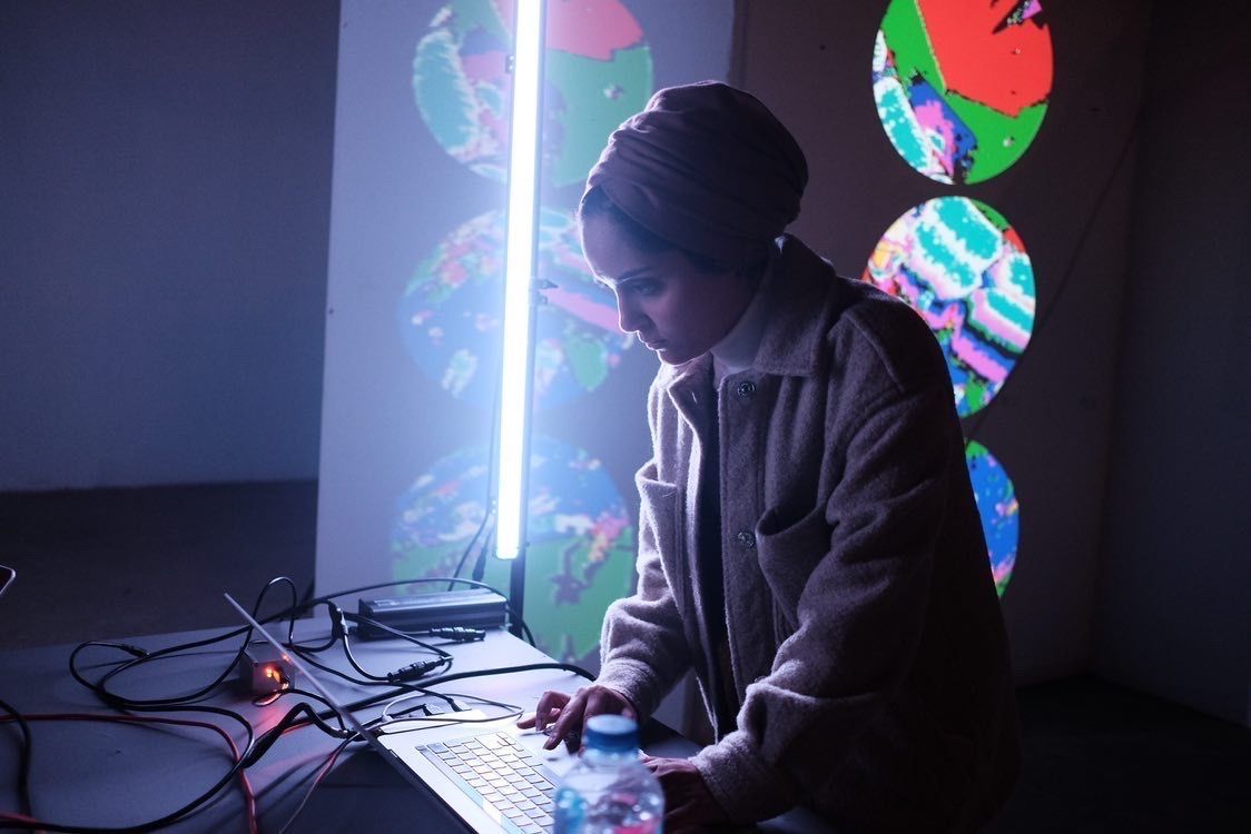 Artist and designer Hadeer Omar stands and leans over a laptop, illuminated from the side by a neon bar of light.