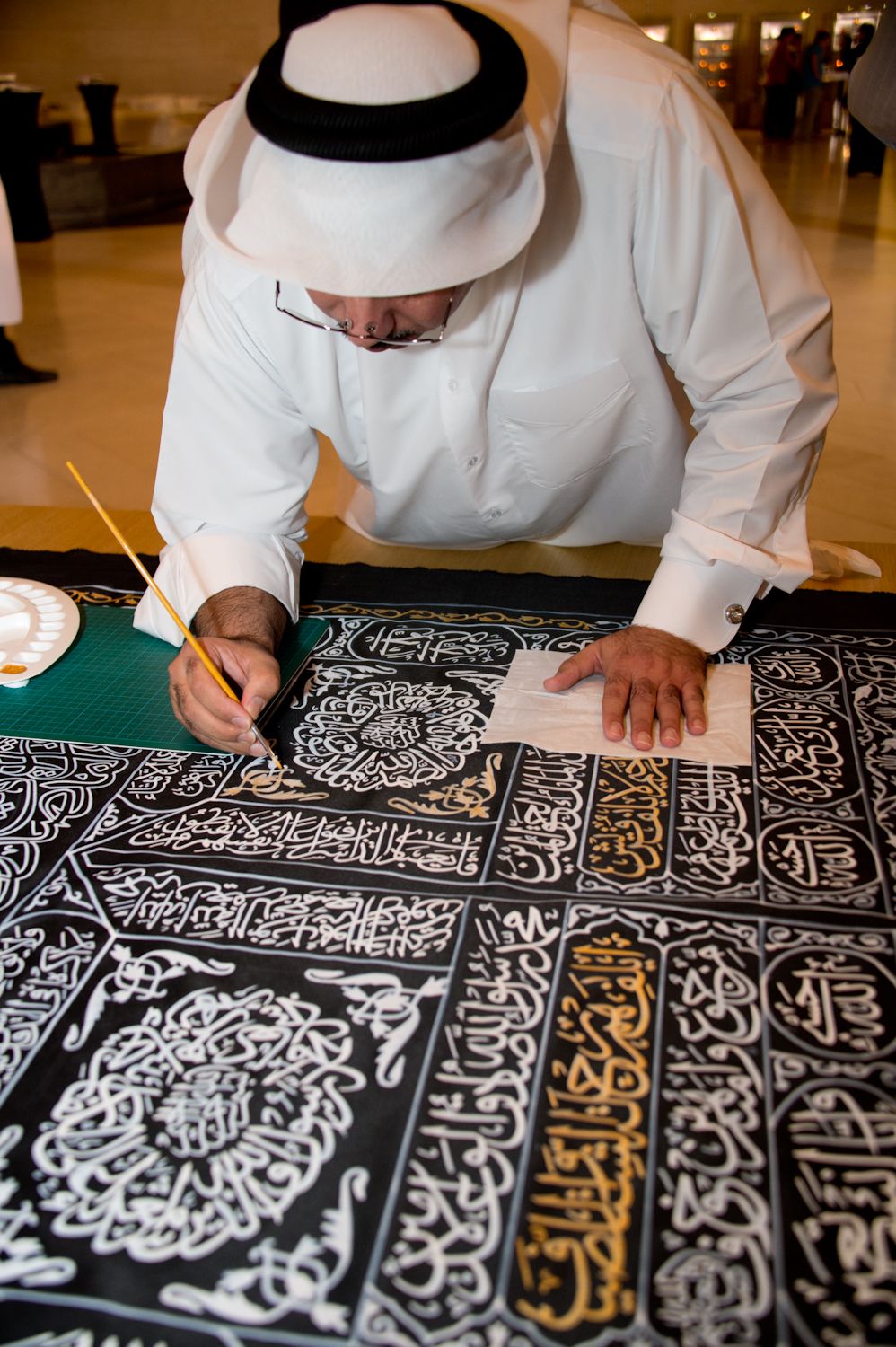 A Middle Eastern artist paints detailed Arabic calligraphy onto a black background with white and gold ink.