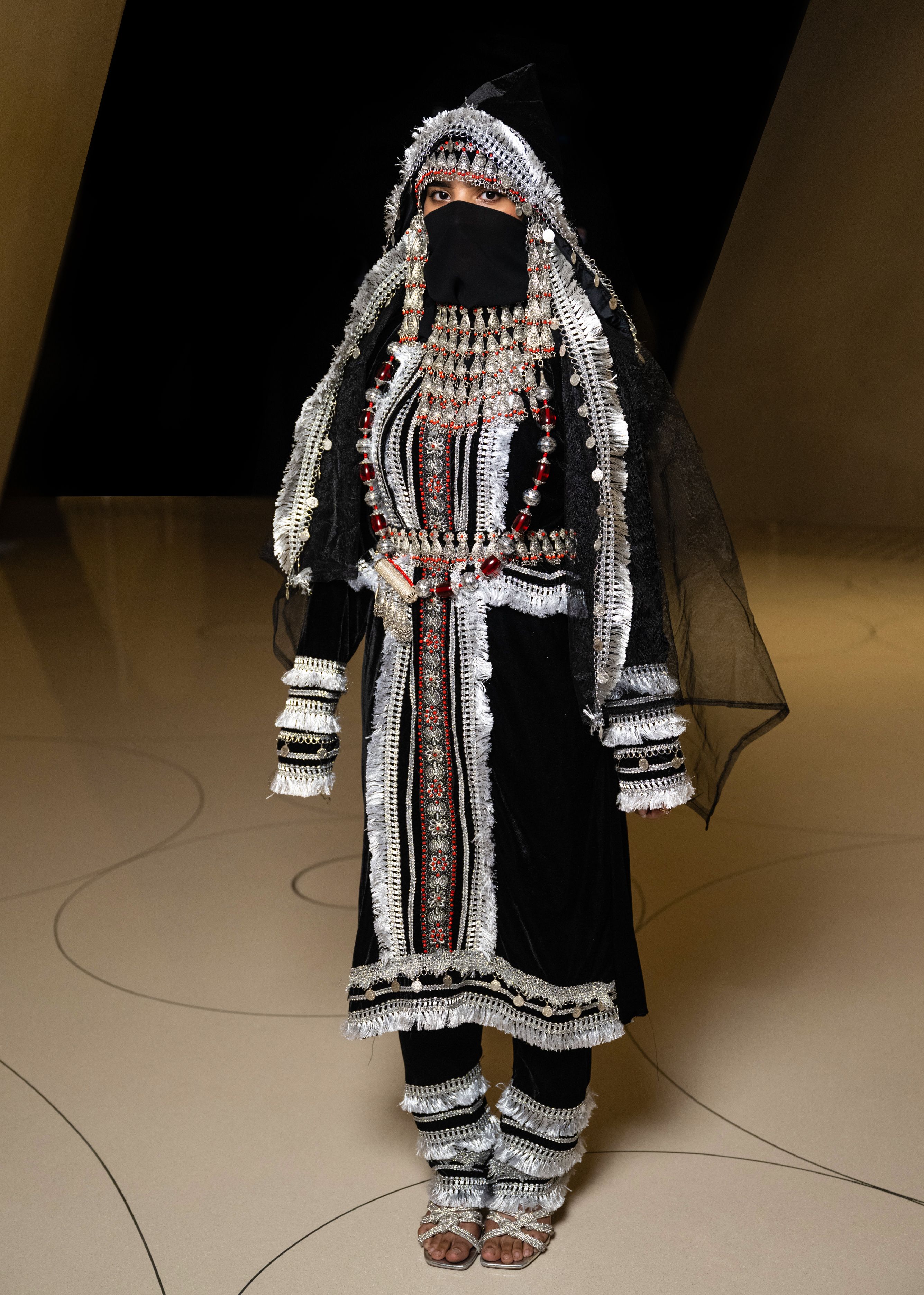 Woman models an embellished black velvet dress and silver embroidered trousers, face veil and head cover.