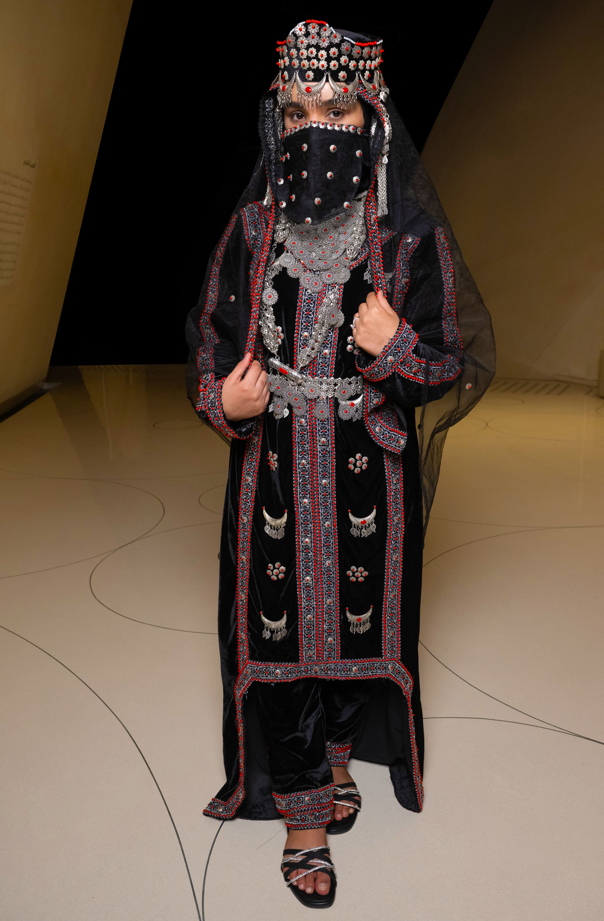 Woman wears a velvet dress embellished with coral and velvet trousers, silver headpiece, ankle bracelet and silver necklace.