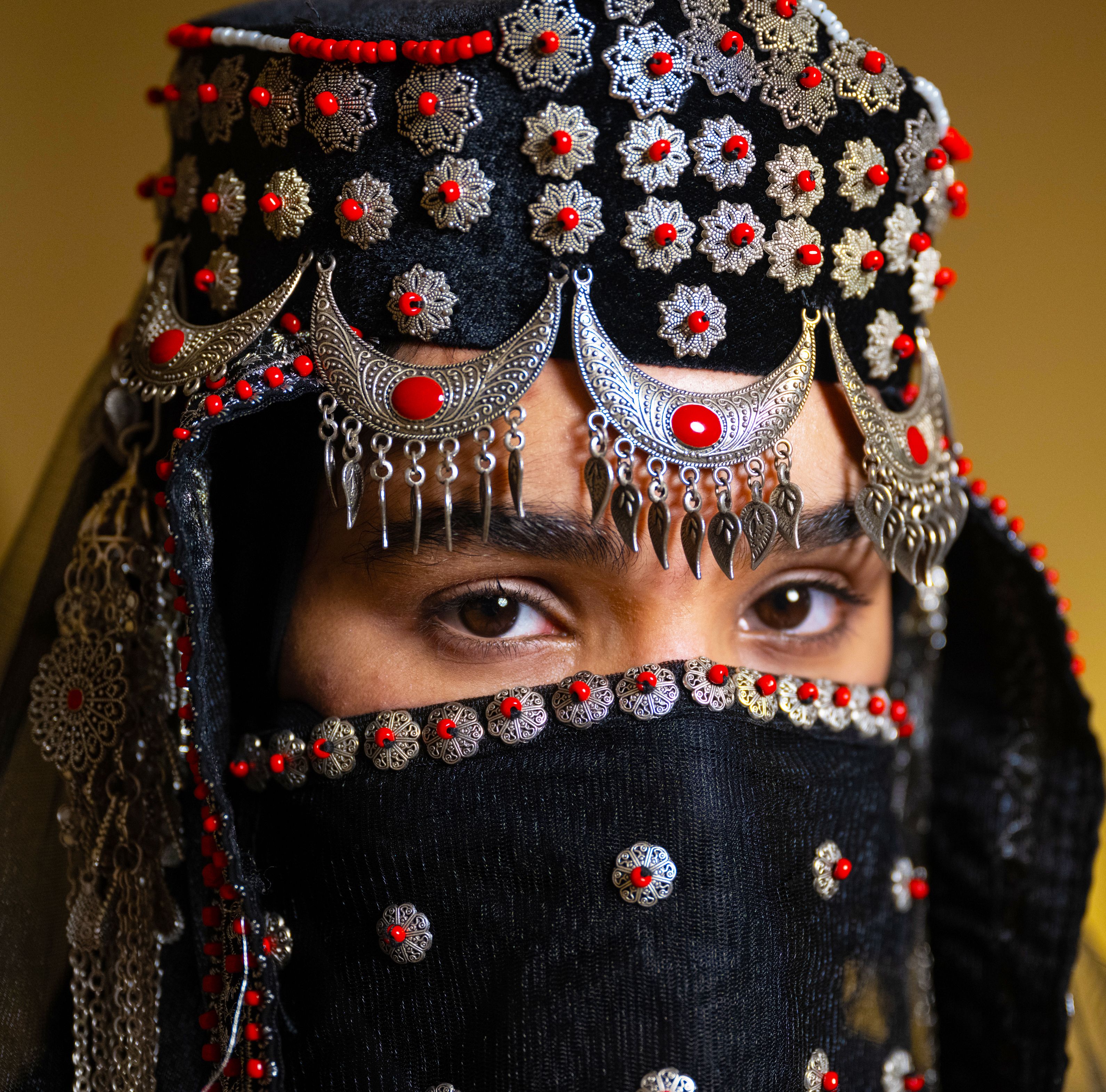 Close up view of a woman's eyes, wearing a traditional Yemeni Maghmuq (veil) and ‘ Asbah (Silver Headpiece) with coral beads.