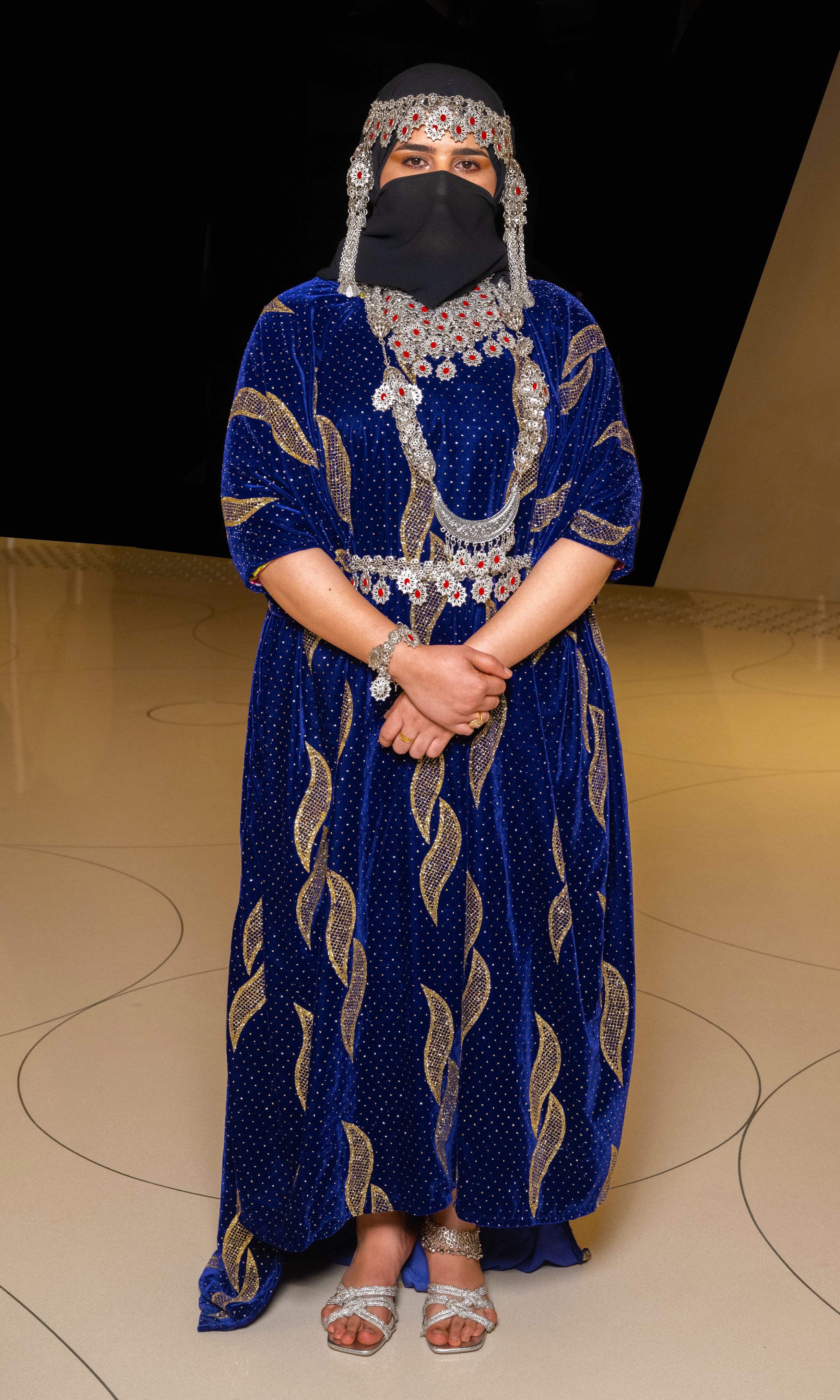 Woman wears a blue dress embroidered with silver threads, silver anklet, necklace and face veil during a Yemeni fashion show.