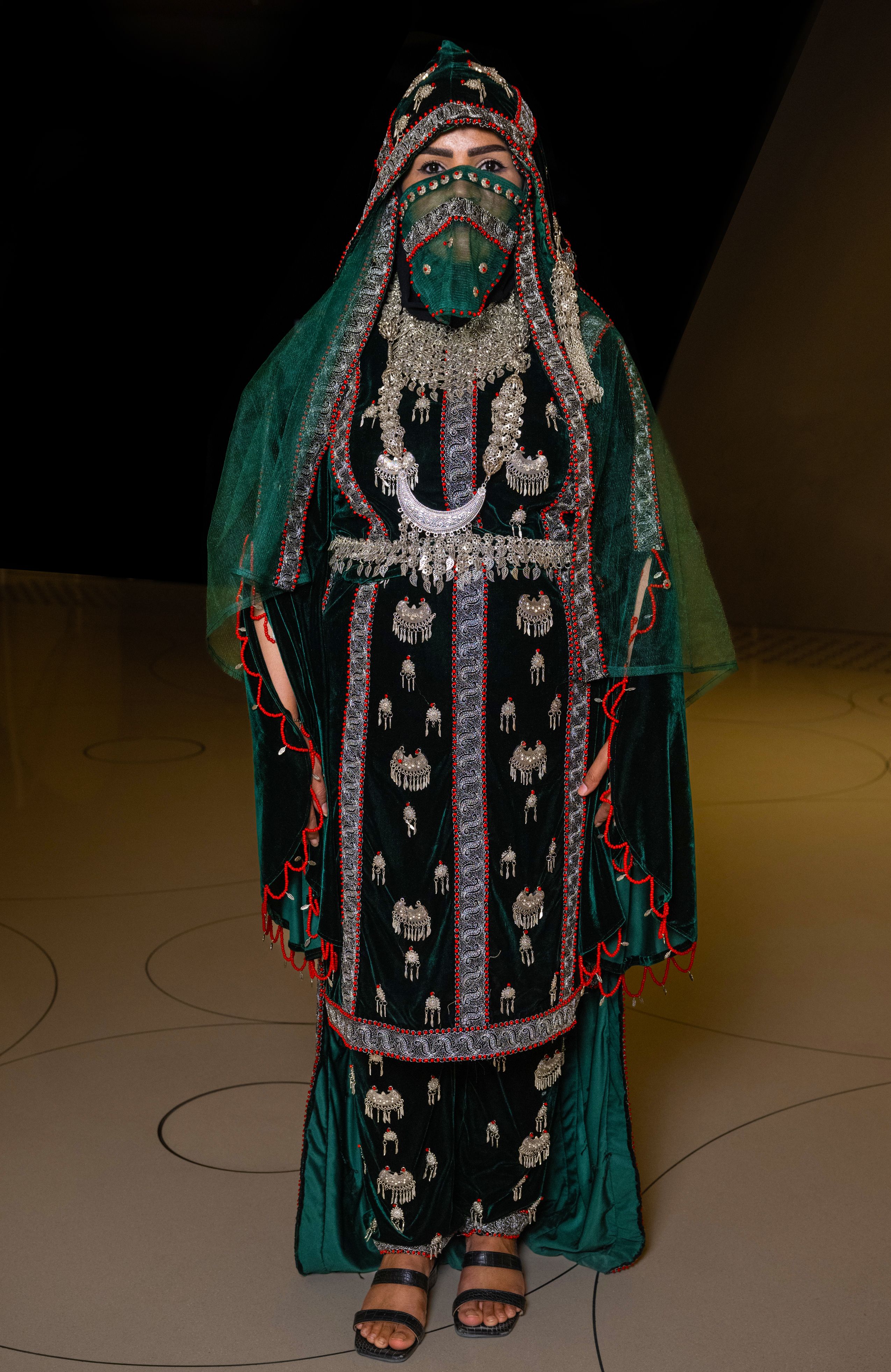 Woman wears a green velvet dress embellished with silver and coral and a head covering traditionally worn in Yemen.