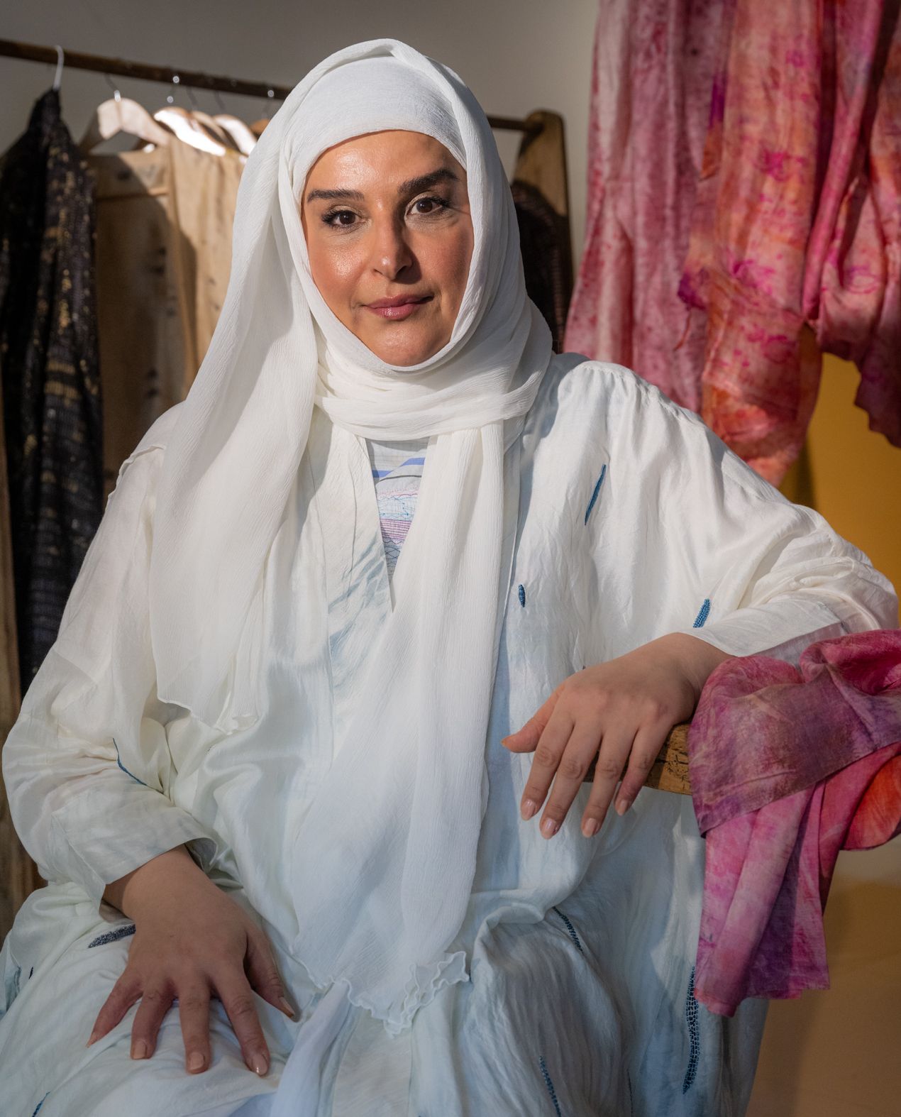 Fashion and textile designer Mashael Alnaimi sits in a chair in front of hanging fabrics, wearing a white outfit and hijab.