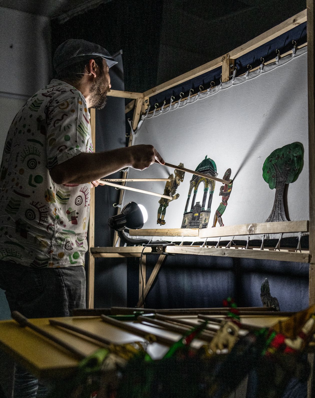 A man performs traditional Turkish shadow puppetry behind a screen, with a puppet on a wooden stick in each hand.