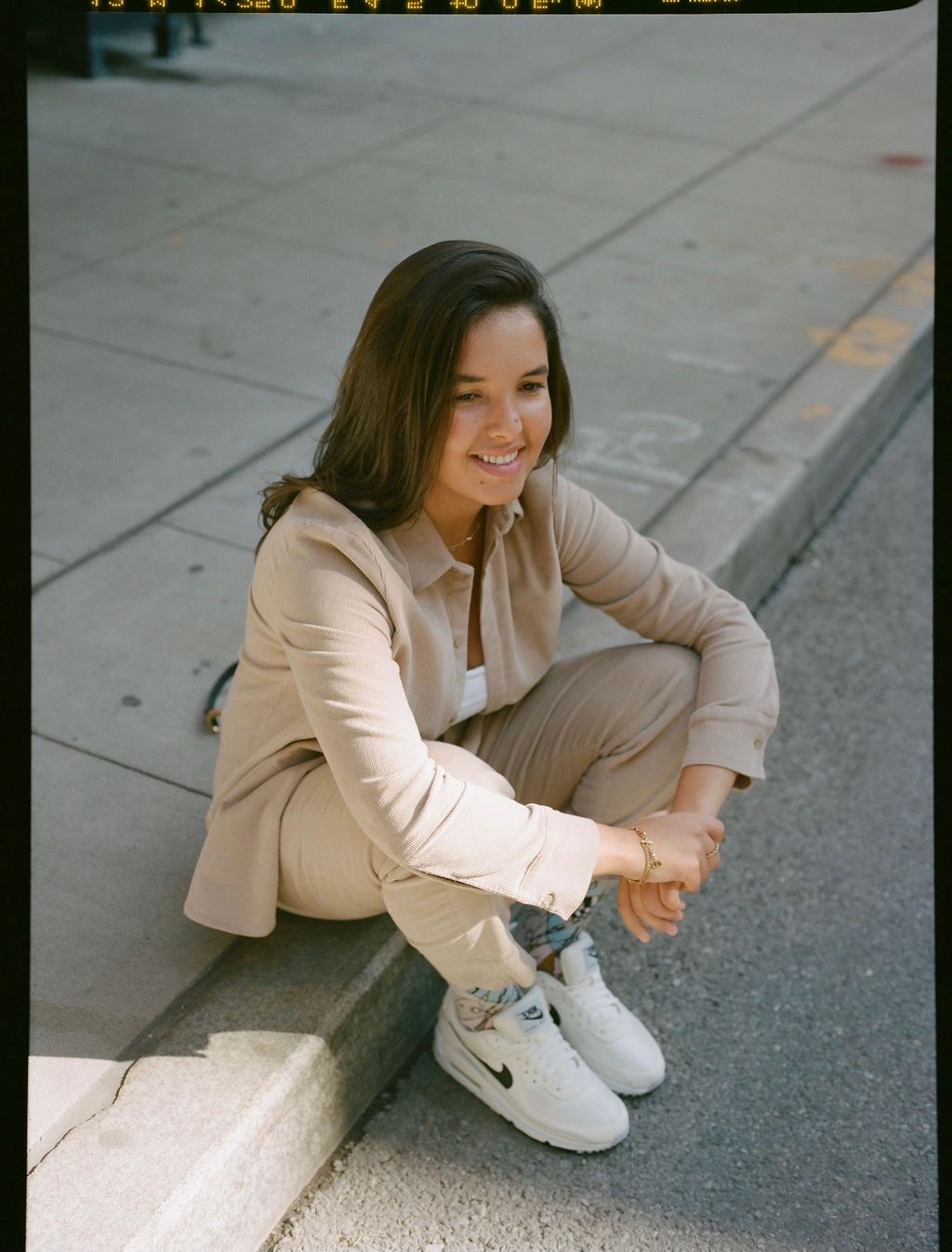 Woman with long dark hair wearing a beige shirt, trousers and sneakers sits on a street kerb 
