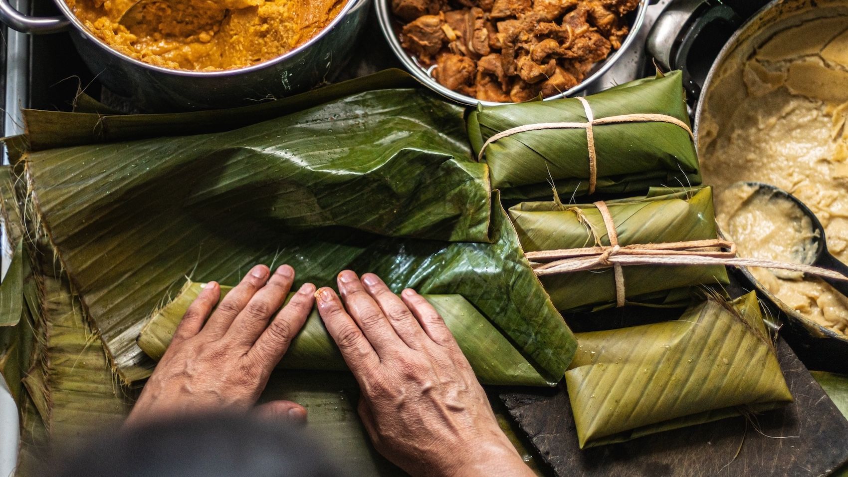Process of making tamales. A man wrapping a corn dough mixture, filled with meat and other ingredients, inside banana leaves before steaming.