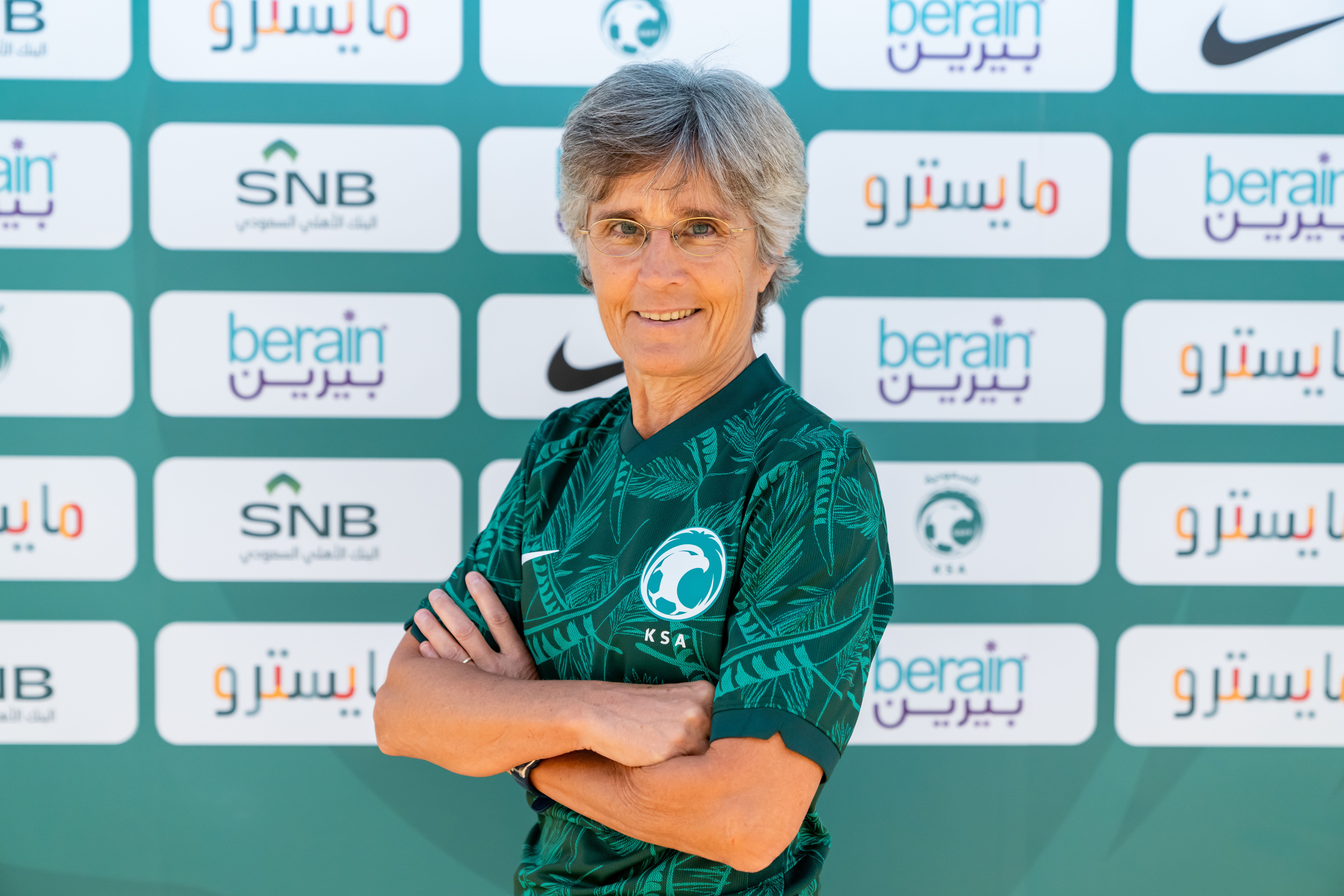 A smiling woman with short grey hair wearing glasses and a green football jersey stands with arms crossed against a backdrop.