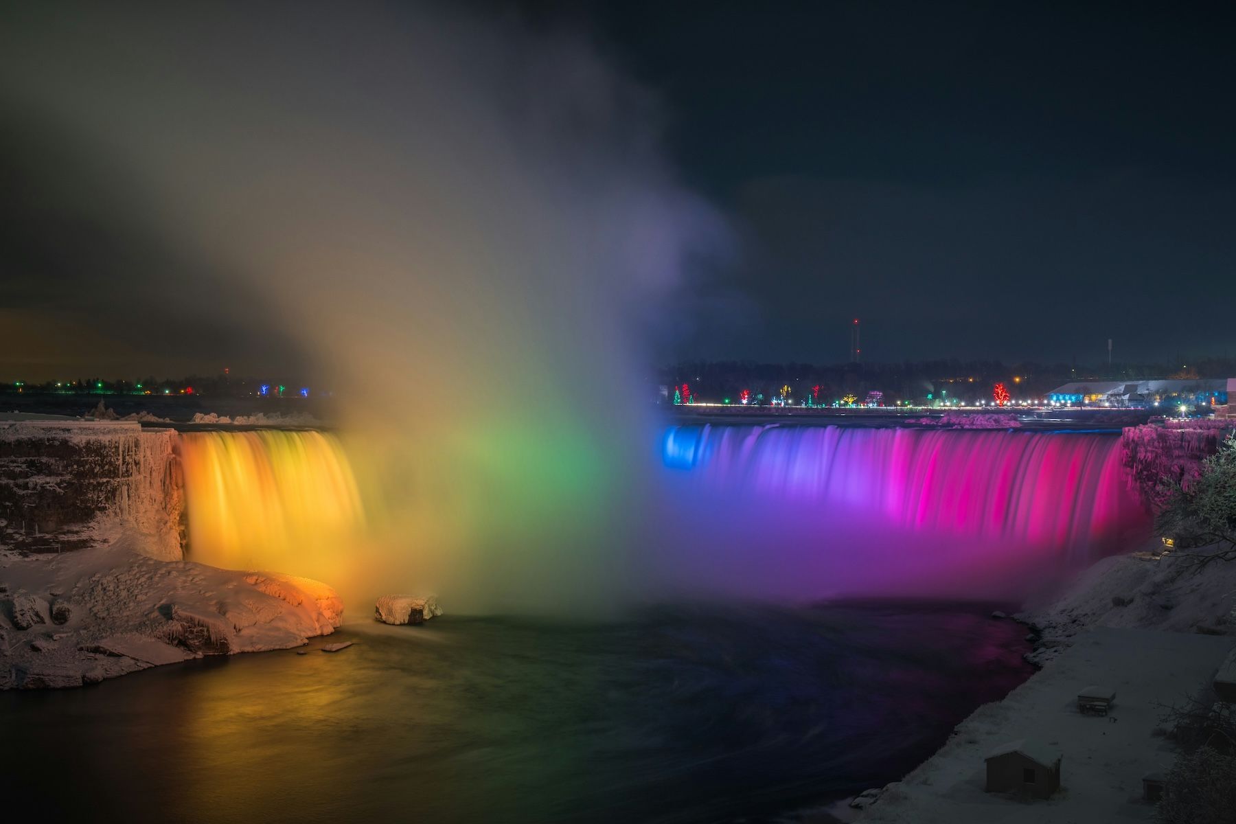 A nightime view of the illuminated Niagara Falls during winter. Falls are lit up by a series of LED lights creating a vibrant display of colours.