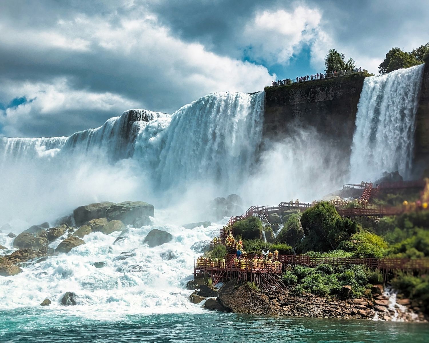 American Falls and Bridal Veil Falls, from Niagara Falls, from the American side. A wooden walkaway, leading to the famous Hurricane Deck and part of the "Cave of the Winds" experience, is clearly visible on the right".