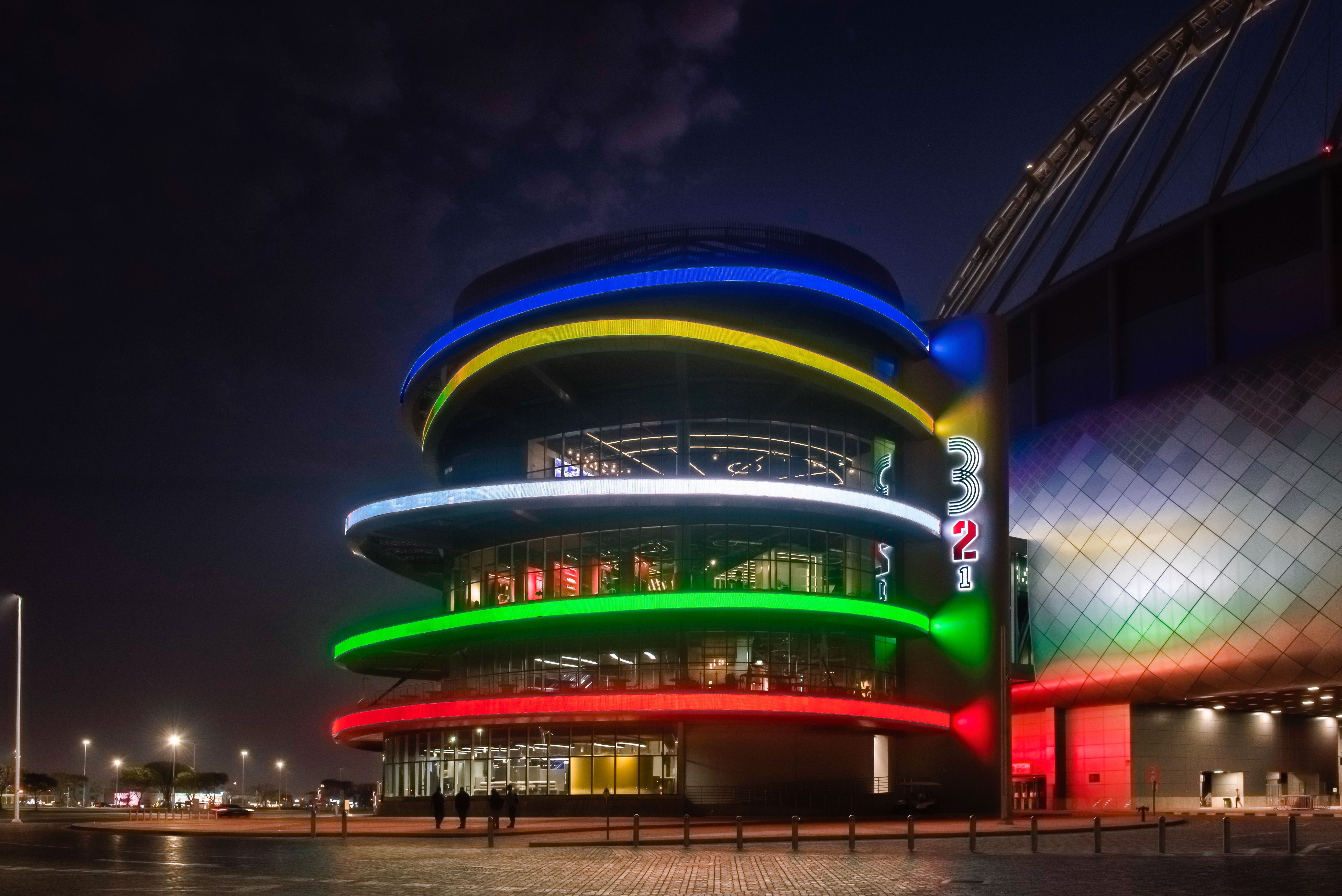 A curved glass building at night, lit with 5 brightly coloured stripes, representing the Olympic Rings. 