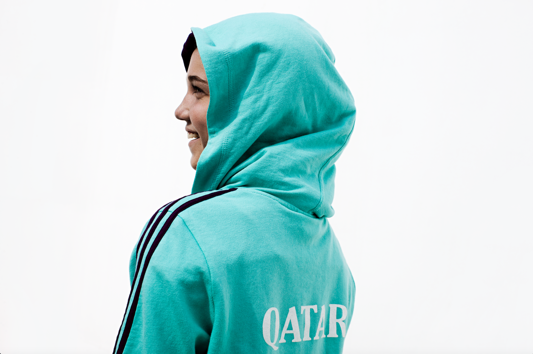 Smiling woman seen from the side, wearing a turquoise hooded sport sweatshirt with Qatar on the back in white text.