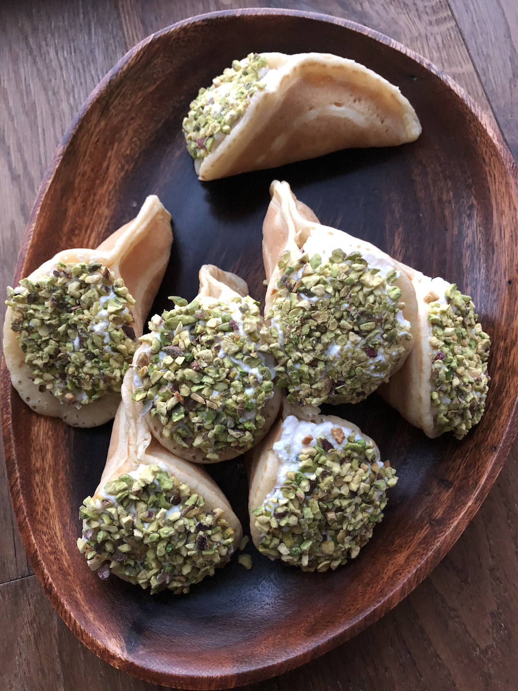 A wooden plate with a pile of atayef pancakes, rolled into a cone with a creamy filling and sprinkled with pistachio nuts.