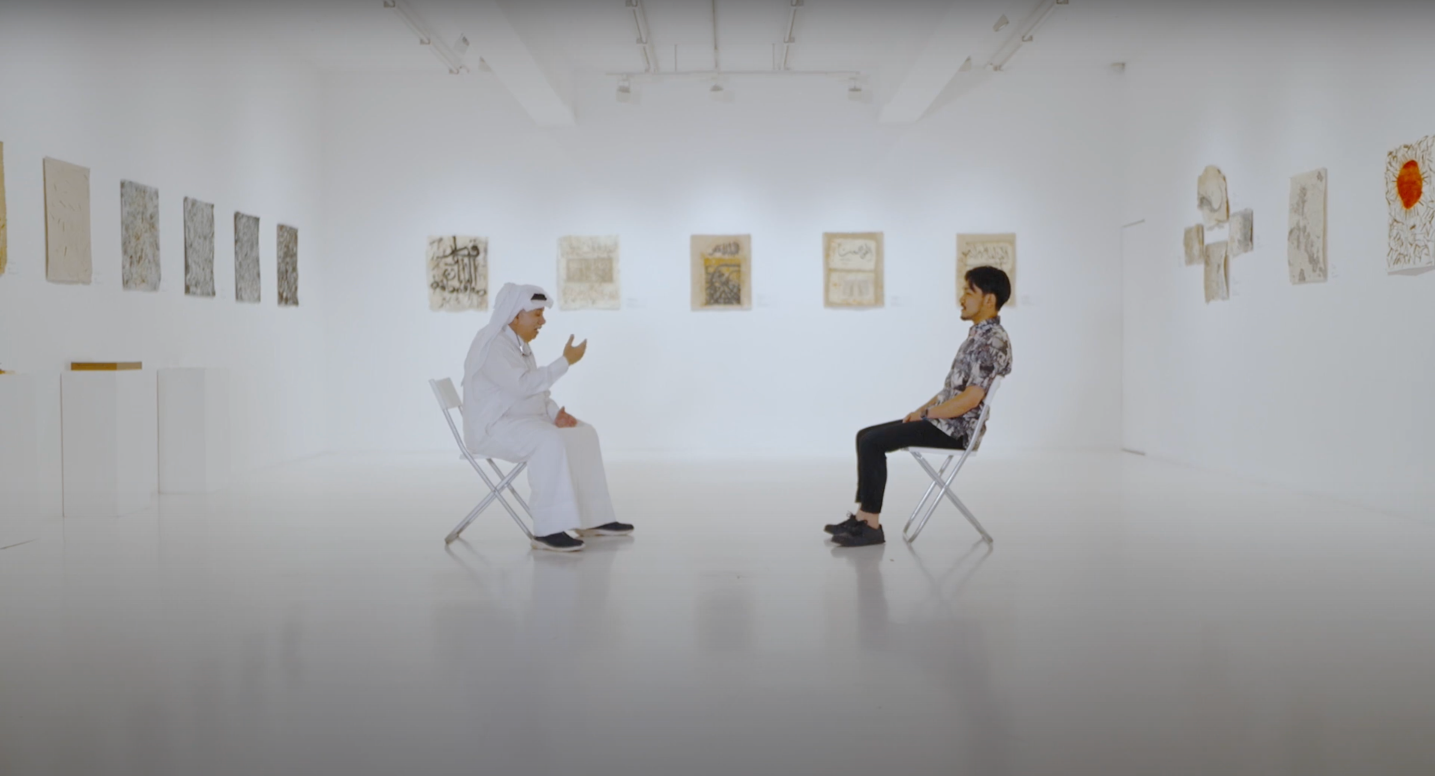 Qatari artist Yousef Ahmad sits on a chair opposite Japanese artist Hayaki Nishigaki in the middle of a white gallery space.