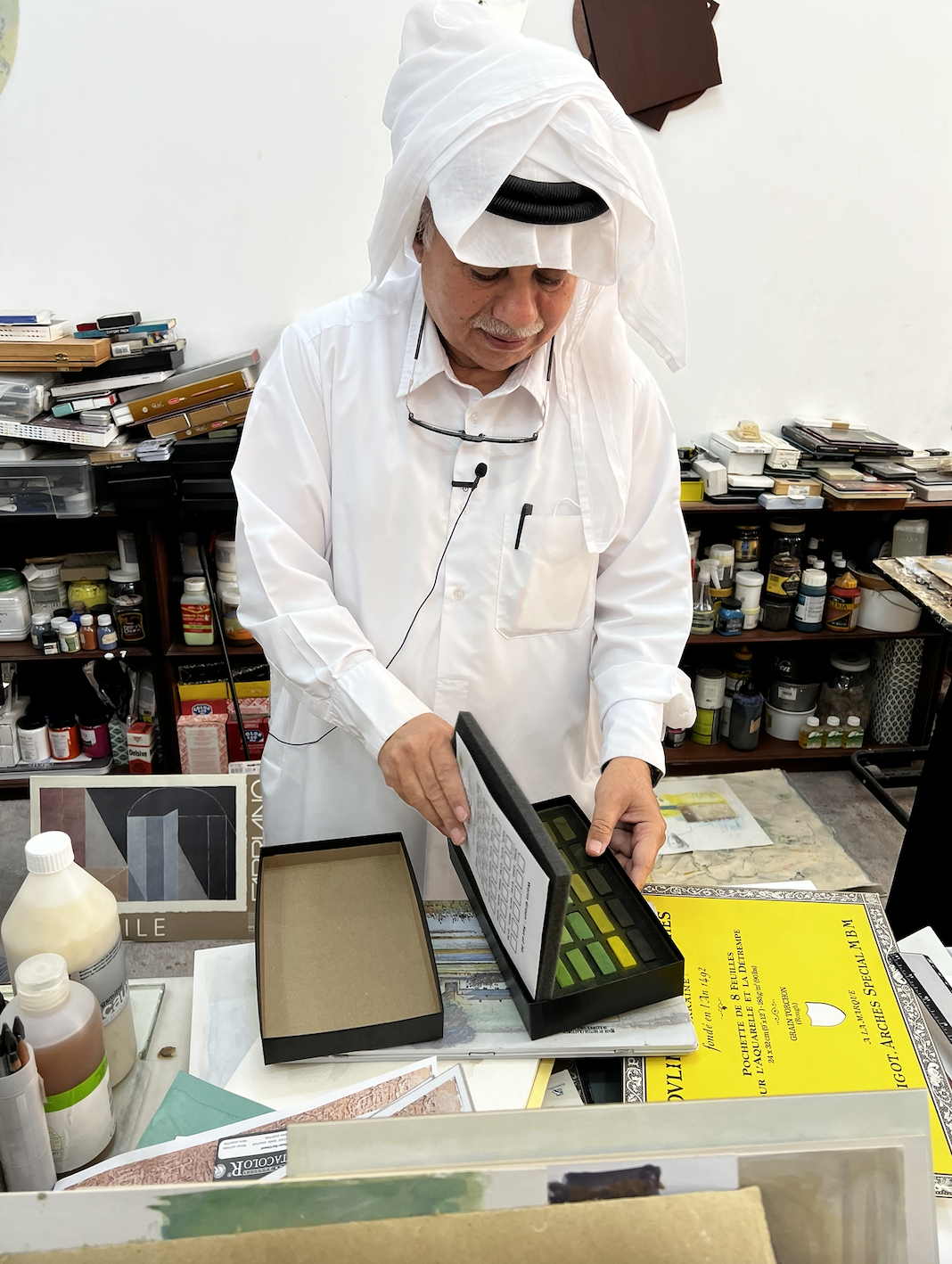A Qatari artist wearing all white traditional clothing looks down at a watercolour paint palate, surrounded by art materials.