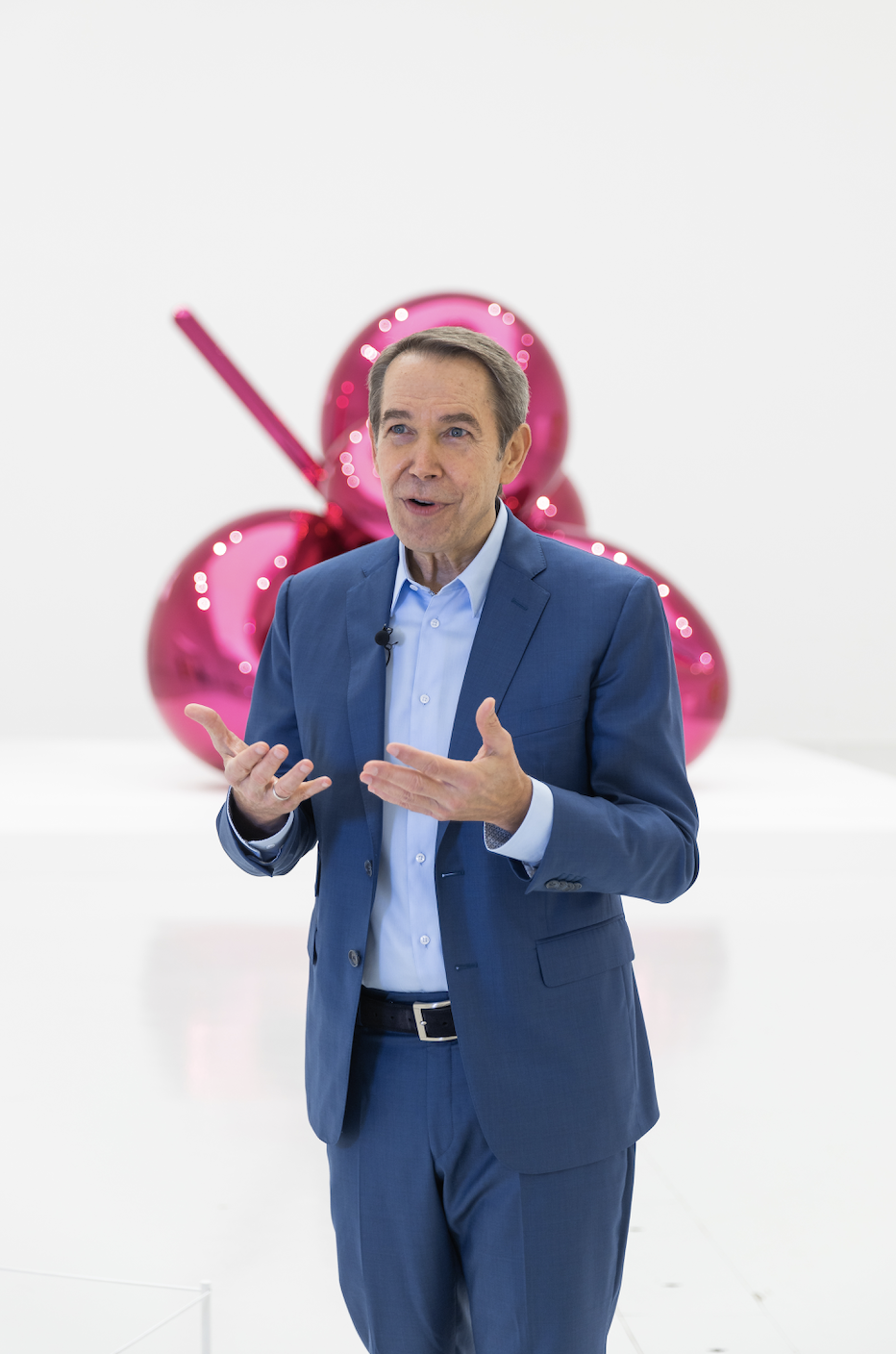 Artist Jeff Koons gives a talk about his exhibition in Doha wearing a blue suit, standing in front of a tall pink sculpture.