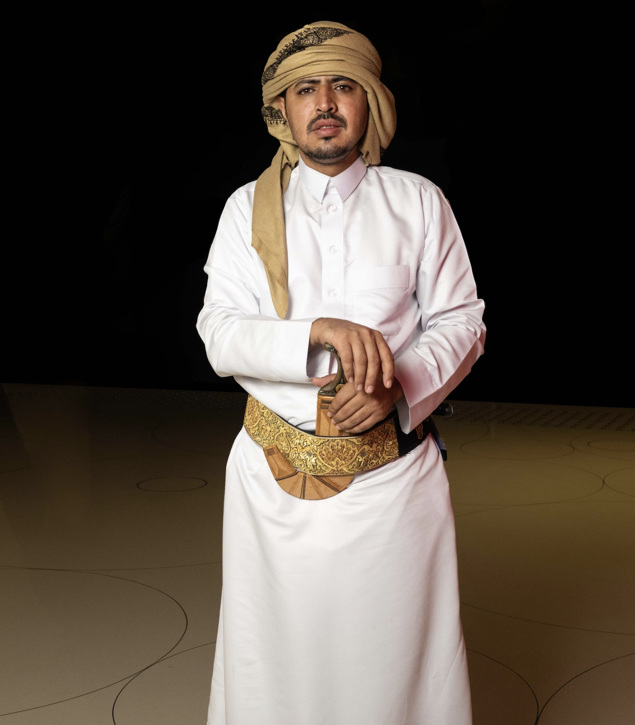 A man models a traditional Yemeni outfit of a long white robe, cloth tied around the head and a Yemeni dagger at his waist.