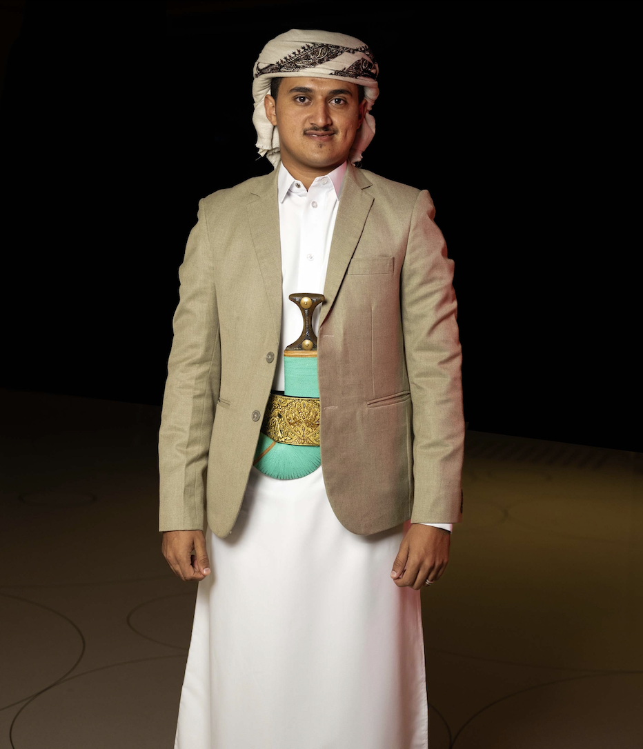 A man wears a traditional Yemeni outfit consisting of a jacket over an ankle length robe and a cloth tied around the head.