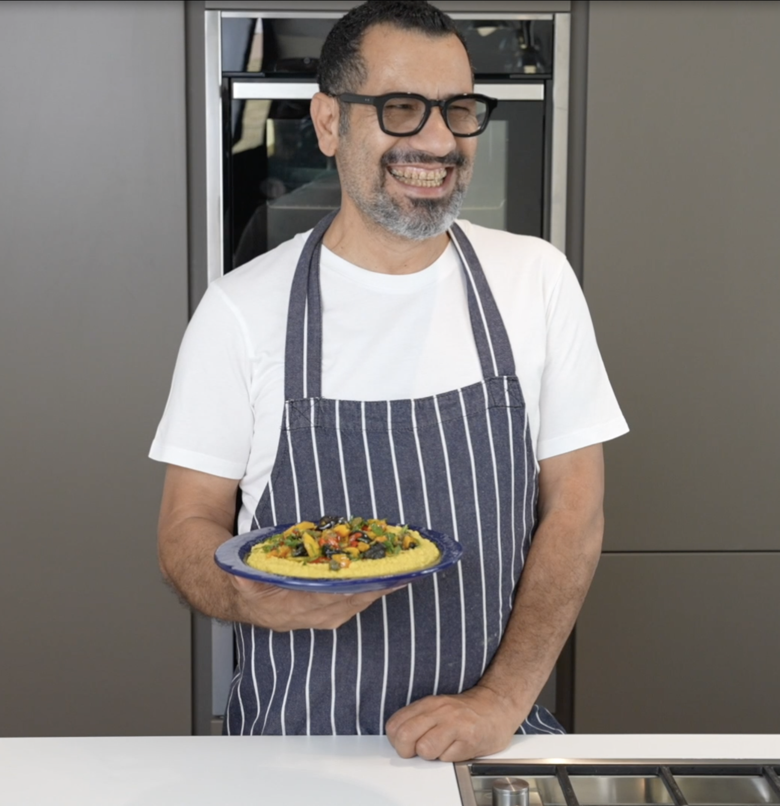 Chef Sami Tamimi wears a white tshirt and grey striped apron, holding a plate of Tunisian chickpeas behind a kitchen counter.