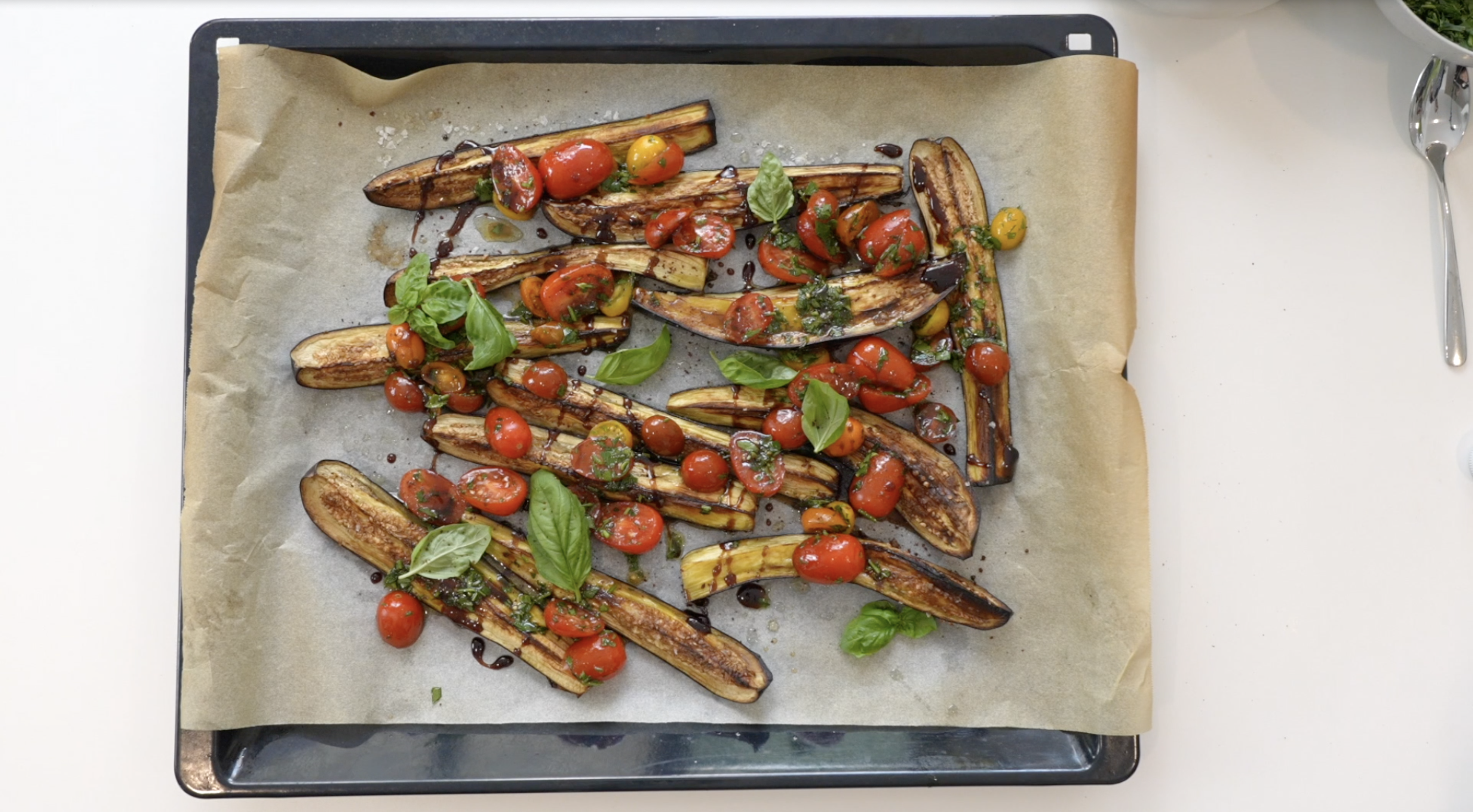 A baking tray with sliced aubergine and halved cherry tomatoes, scattered with basil leaves and drizzled with balsamic vinegar.