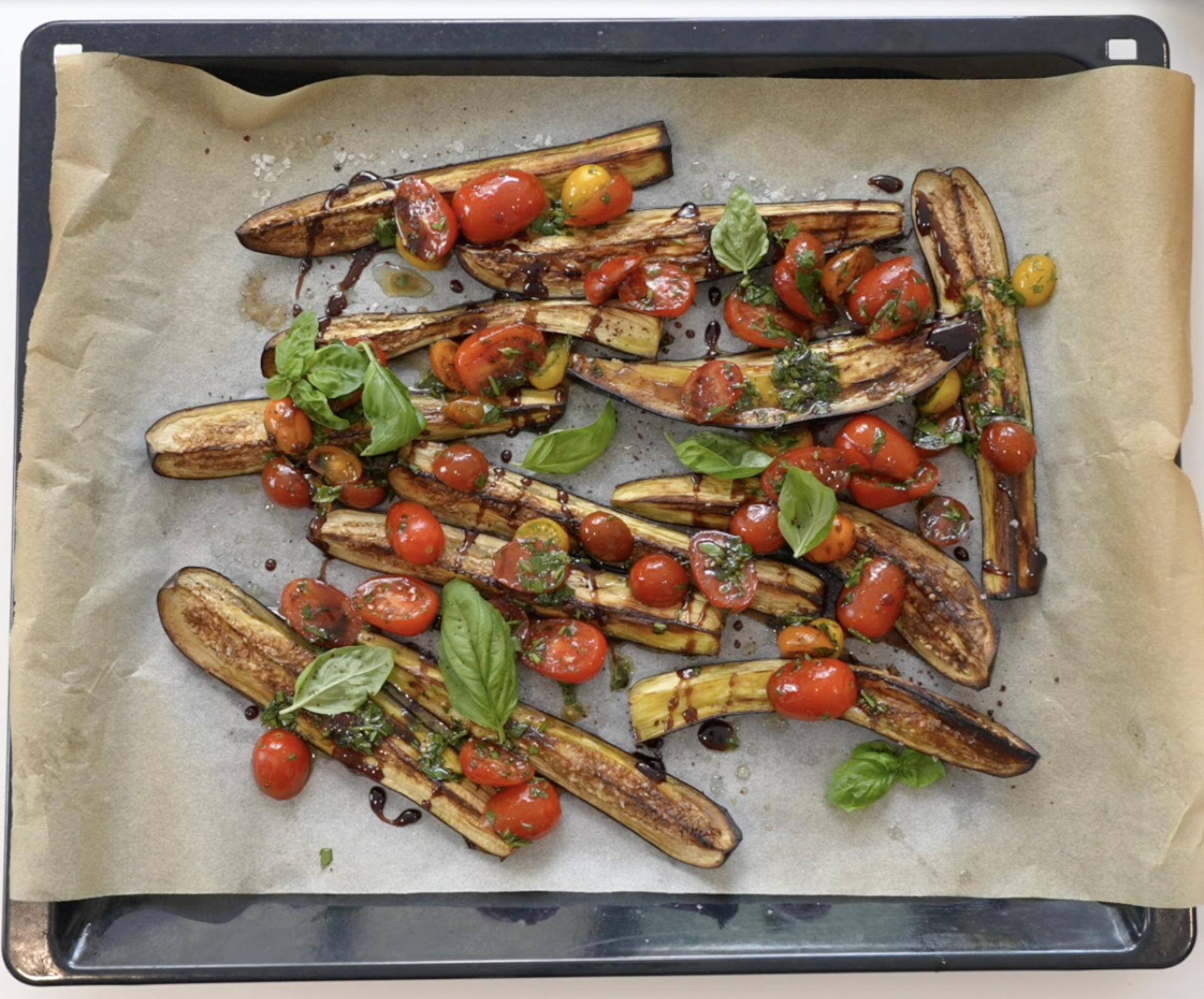 Baking tray with sliced aubergine and halved cherry tomatoes, scattered with basil leaves and drizzled with balsamic vinegar.