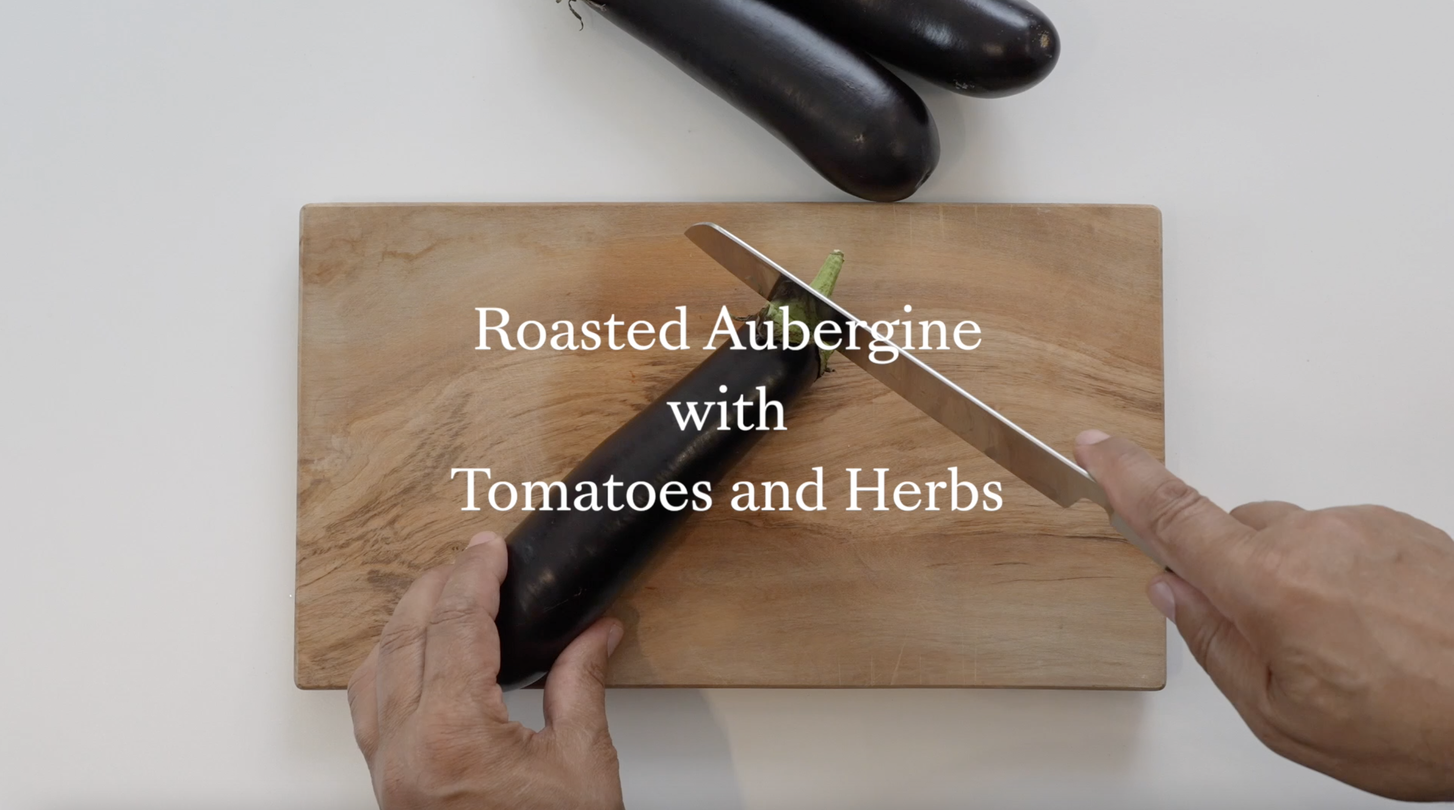 Two hands seen from above slice an aubergine with a knife on a wooden chopping board.