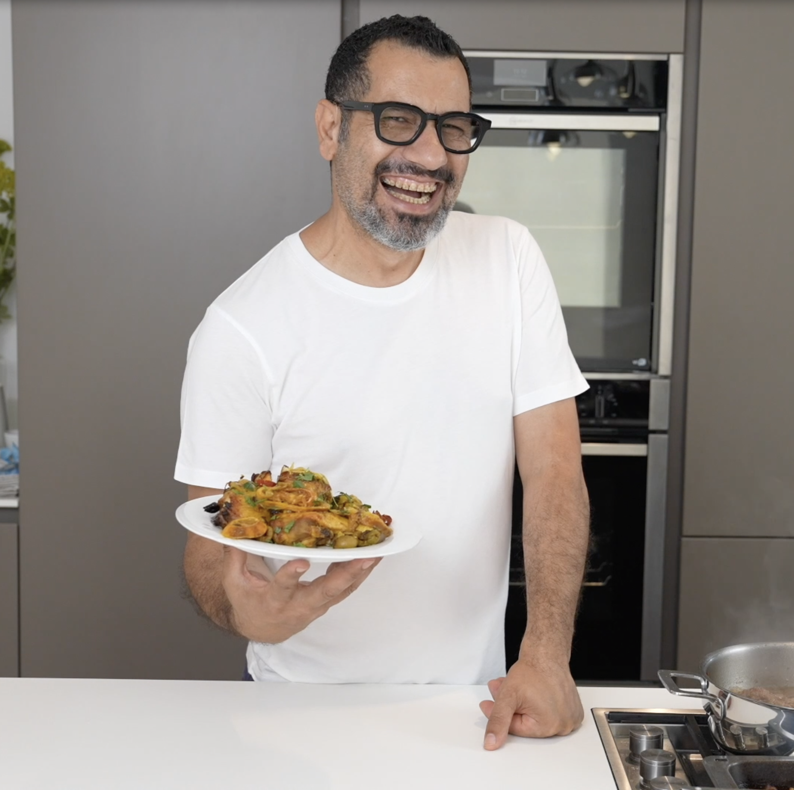 A chef wearing a white t-shirt and glasses smiles while holding a plate of moroccan chicken, standing at a kitchen counter.