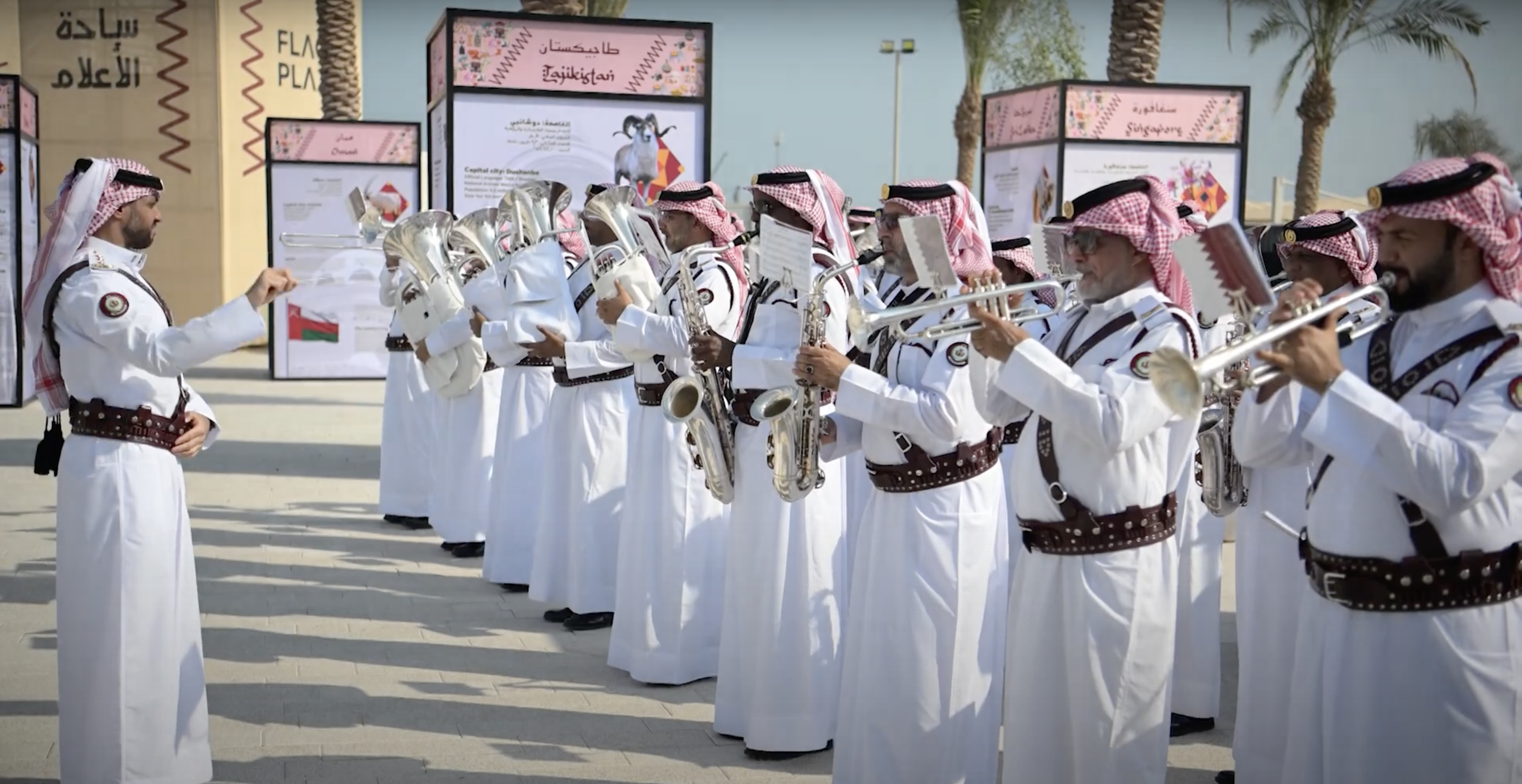 A band wearing traditional Qatari dress play brass instruments outside in the sunshine.