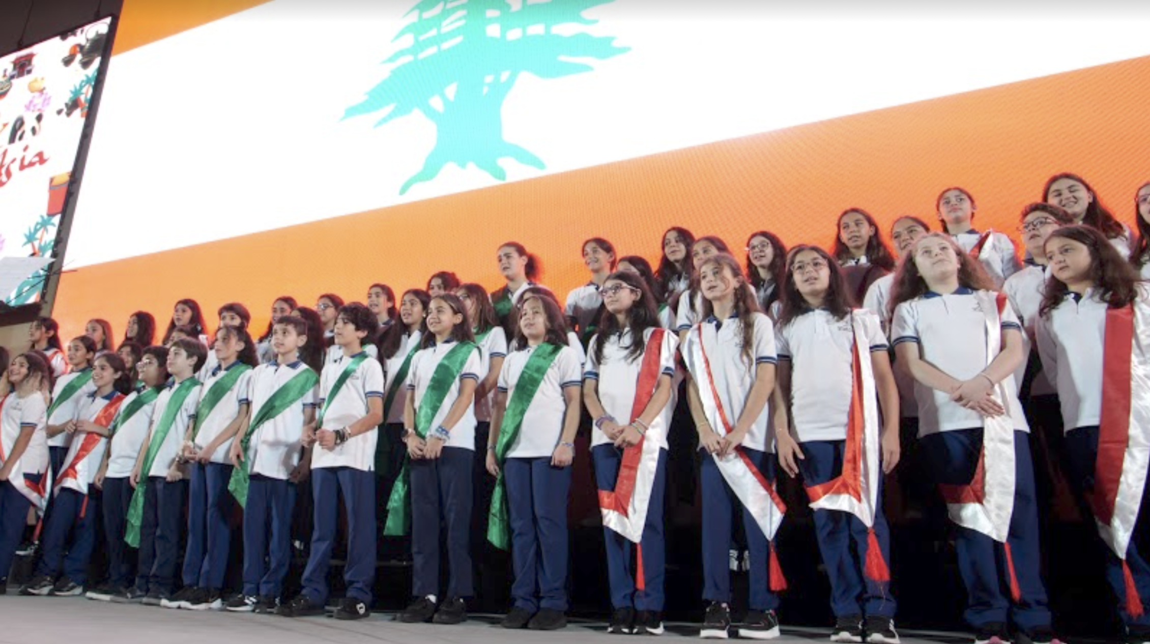 A choir of children wearing red and green sashes perform a song on stage in front of a giant Lebanese flag.