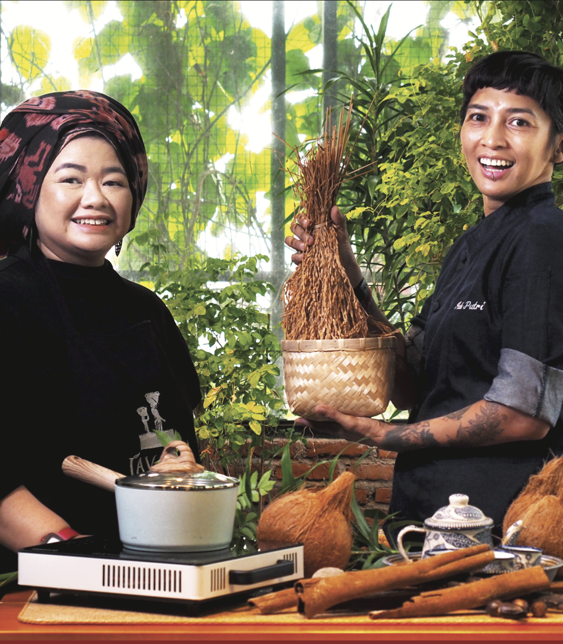 Two women wearing chef jackets prepare food with Indonesian ingredients in an outdoor kitchen.