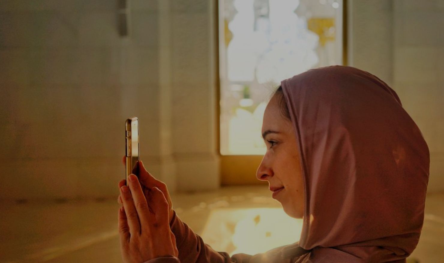 Middle Eastern woman wearing a pale pink hijab takes a photograph using a smartphone.