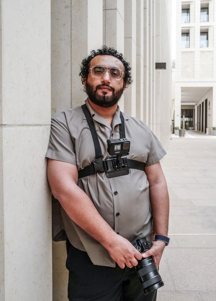 Photographer Ammar Alqamash wearing a grey short sleeved shirt and sunglasses, holding camera equipment.