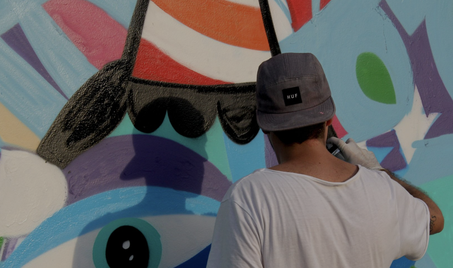 A man wearing a backwards cap and white t-shirt paints a colourful mural with a can of spray paint in his hand. 