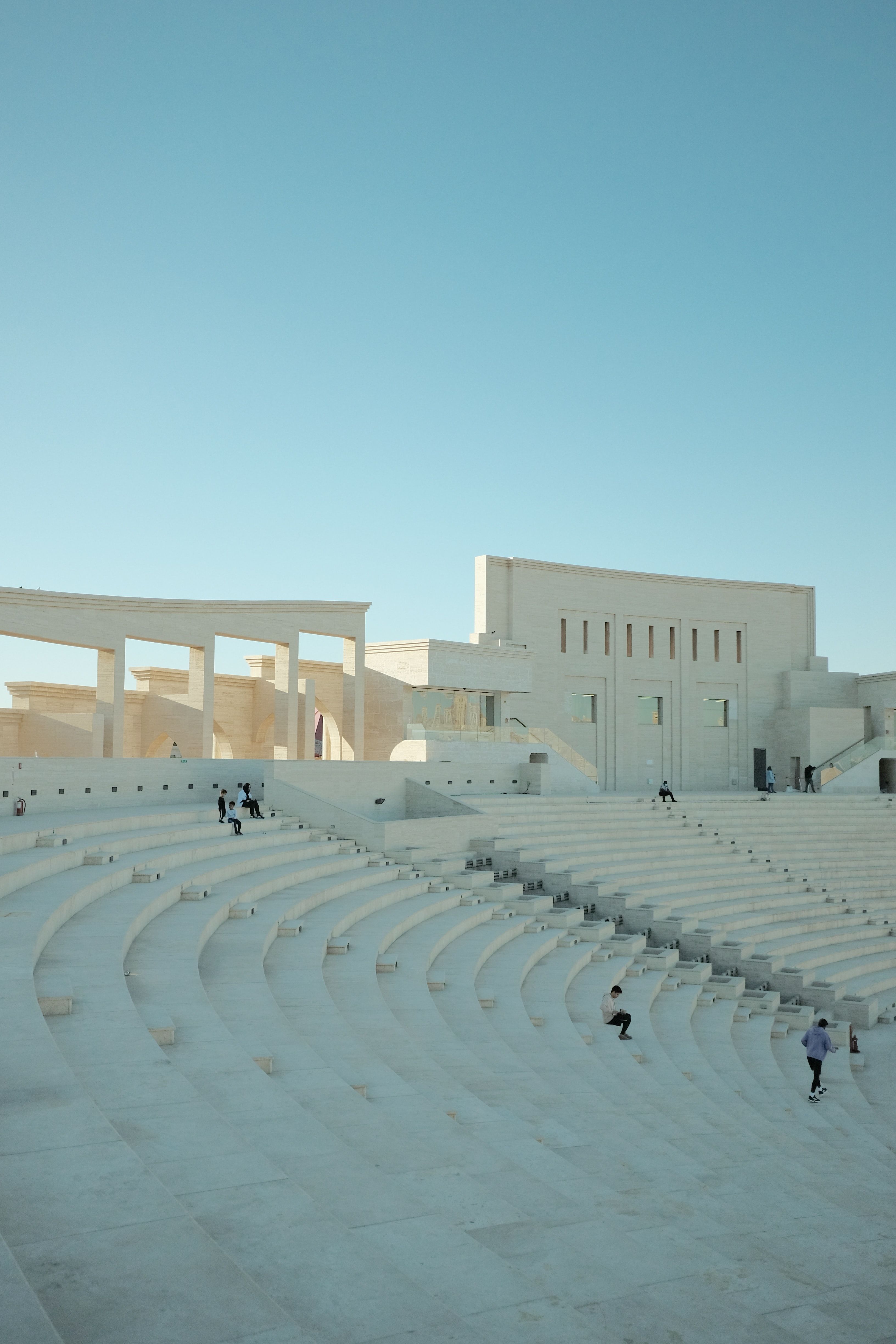 A white stone outdoor amphitheatre at Katara Village with tiered seating, under a clear blue sky.